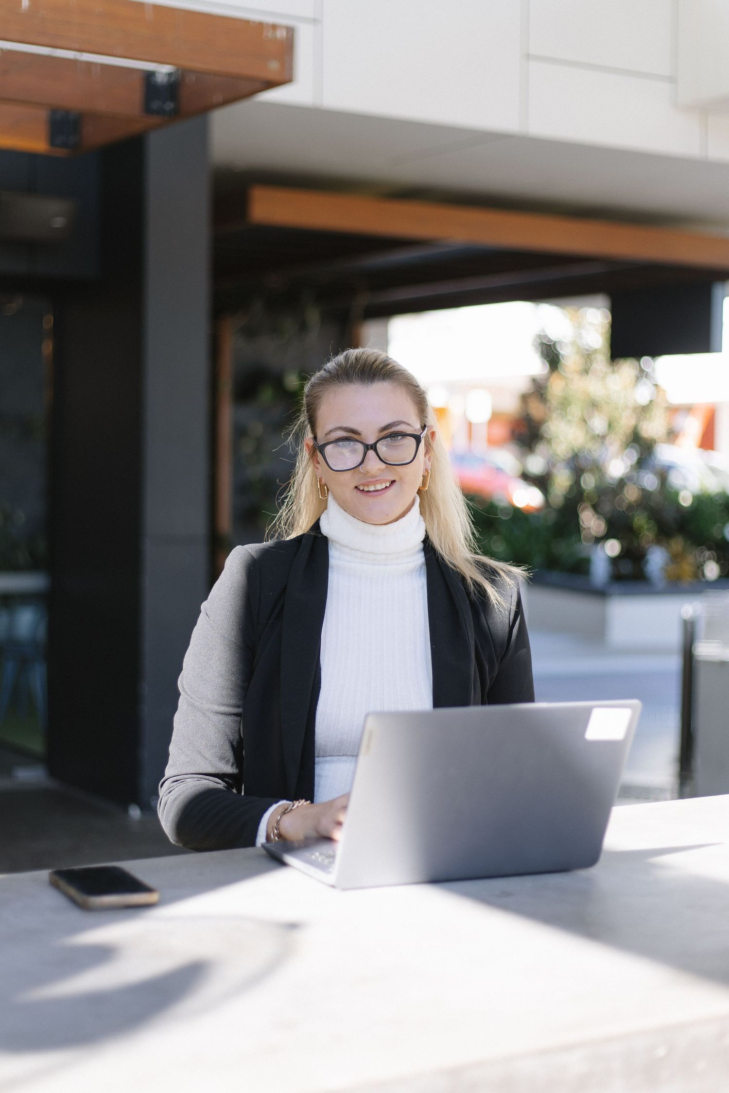 A person in business attire works on a laptop at an outdoor table with modern architecture in background.
