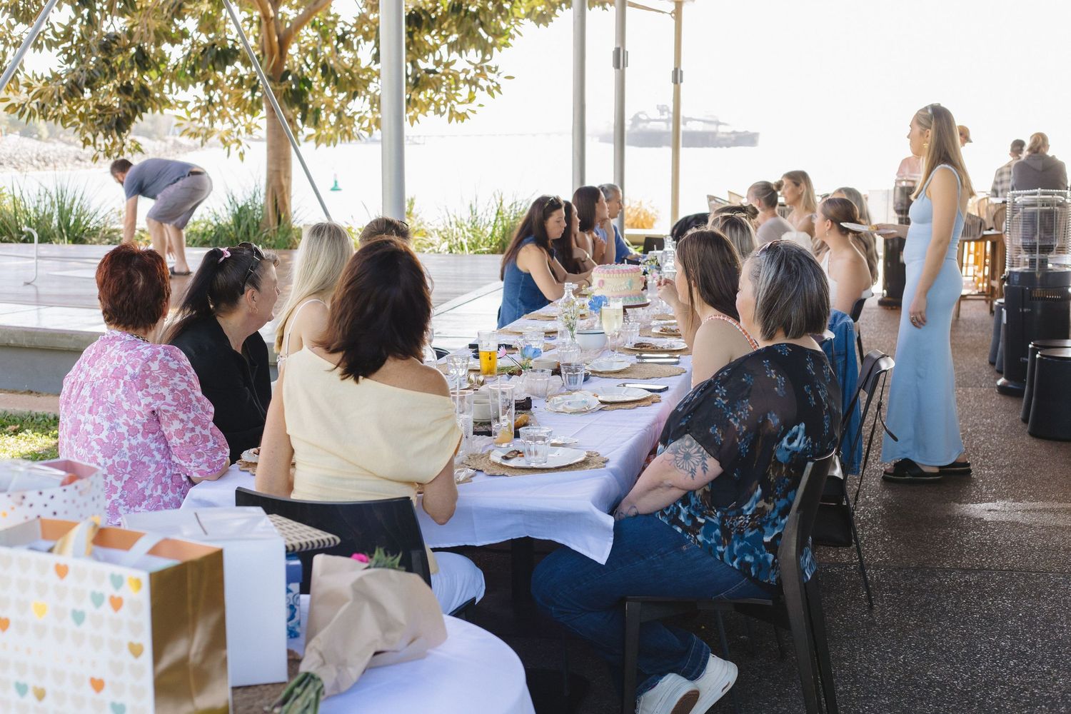 Outdoor party setup with guests seated at long tables under a covered patio area.