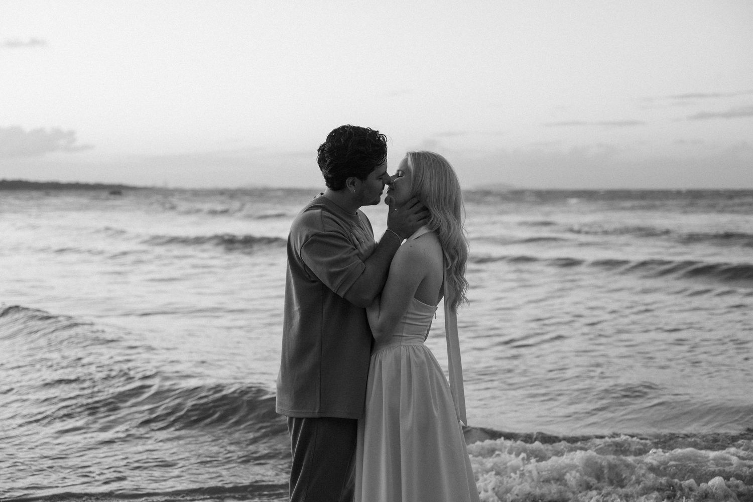 Black and white photo of a couple sharing a romantic moment on a beach at sunset with ocean waves behind them.