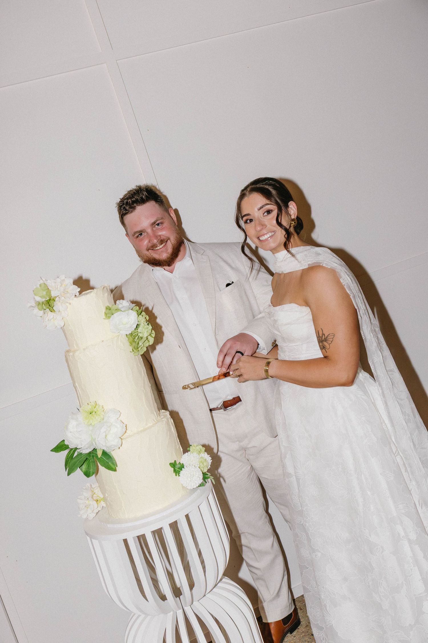 Elegant white wedding cake with floral decorations being cut by a couple in formal attire.