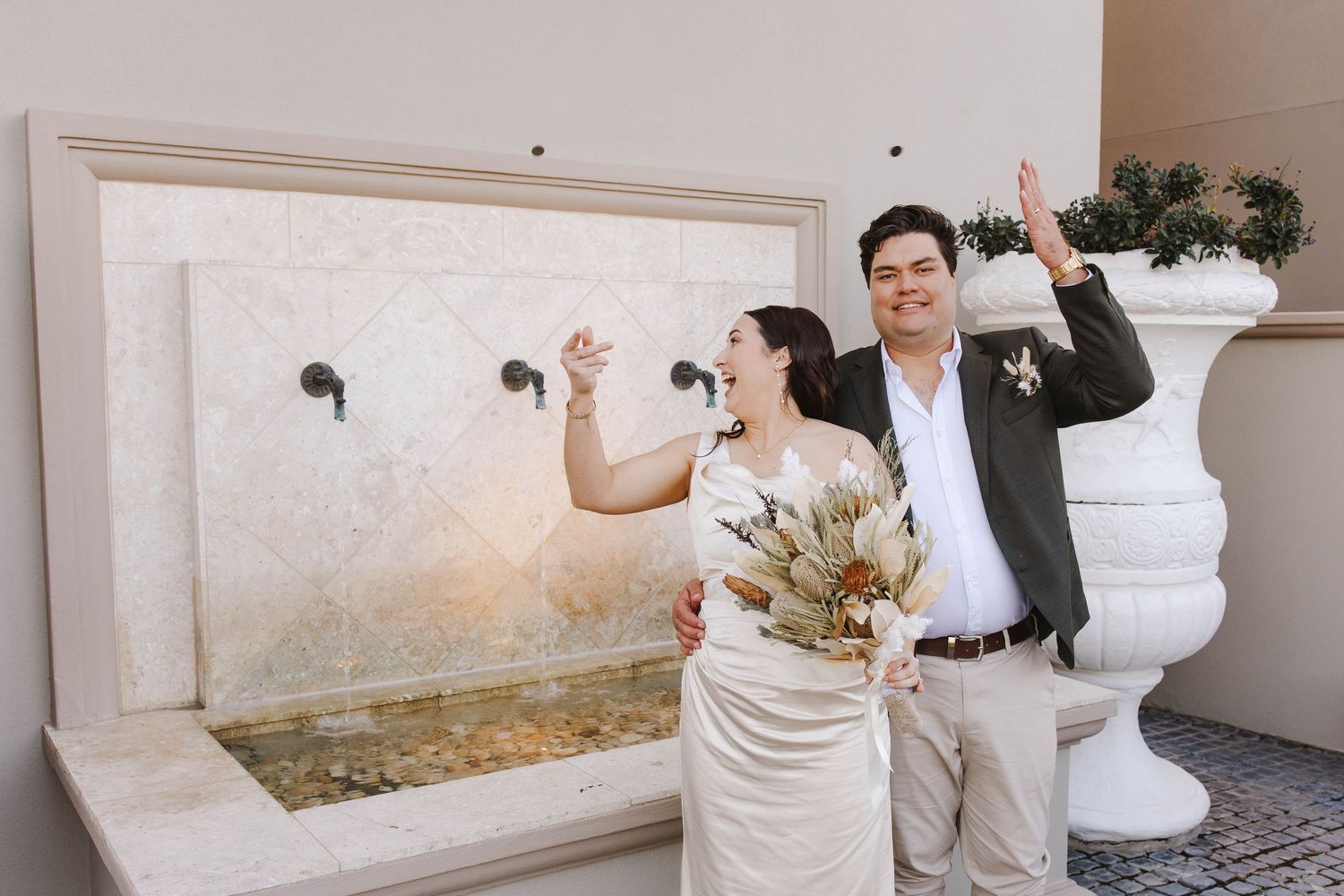 Joyful newlyweds celebrate near a stone fountain, bride holding a boho bouquet and groom in a gray suit, both raising hands.