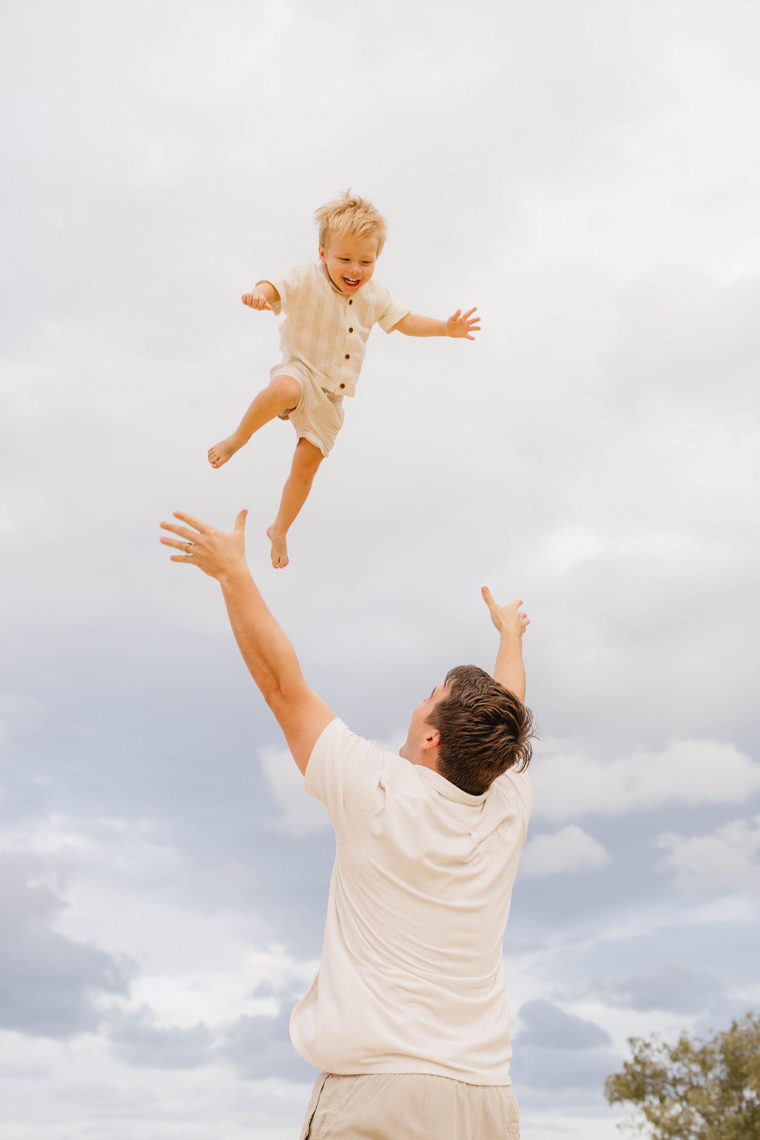 Joyful parent tossing child in the air against cloudy sky background.