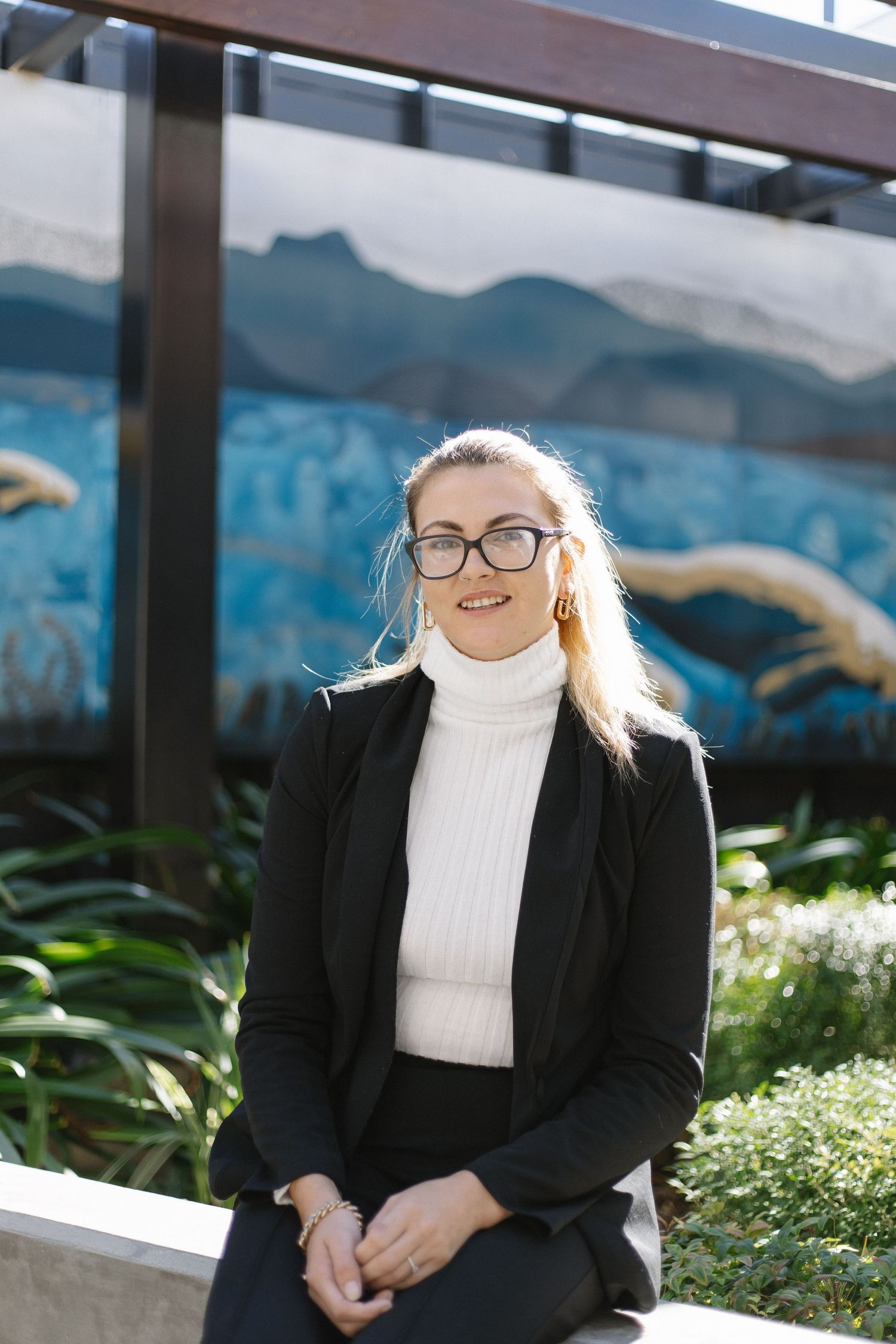 Professional in black blazer and white turtleneck seated outdoors with mountain backdrop.