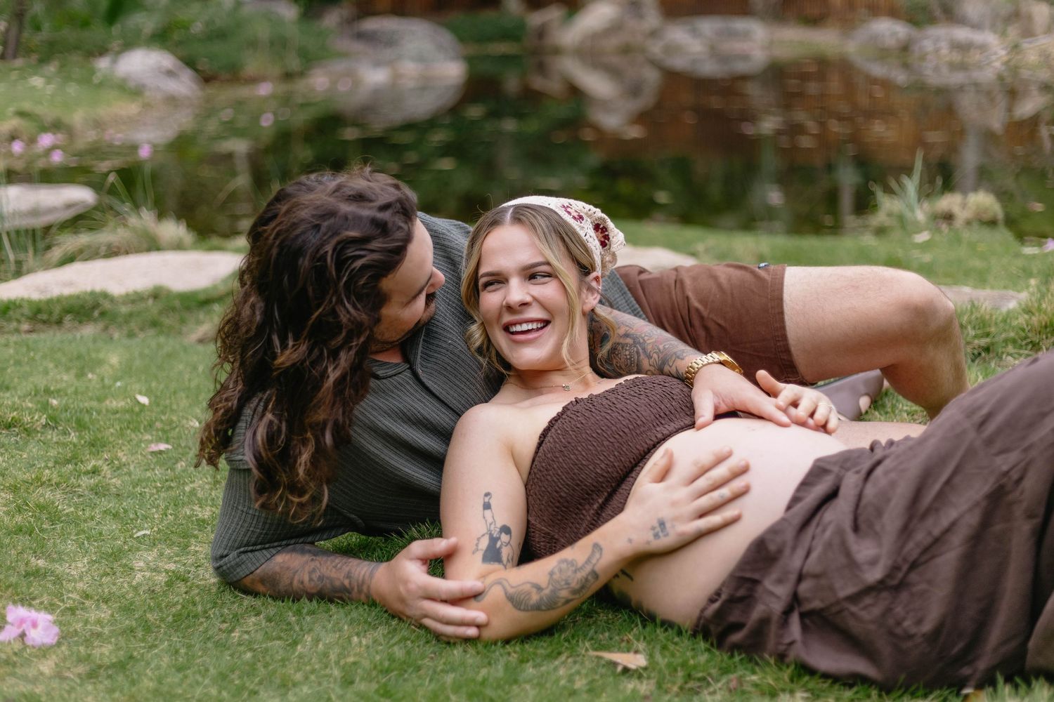 Couple poses playfully on grass with purple flowers in garden setting.