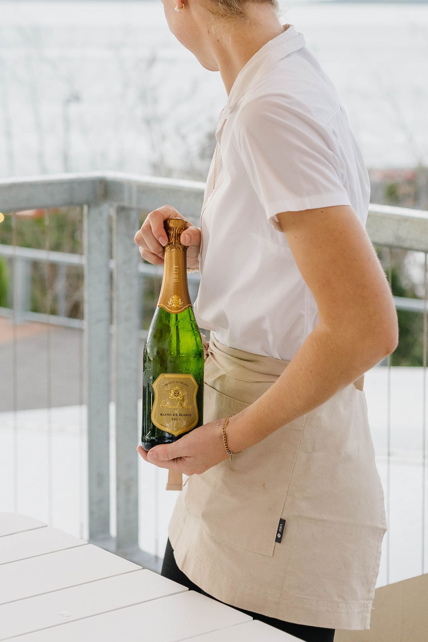 Server in white uniform and beige apron holding champagne bottles on outdoor terrace.