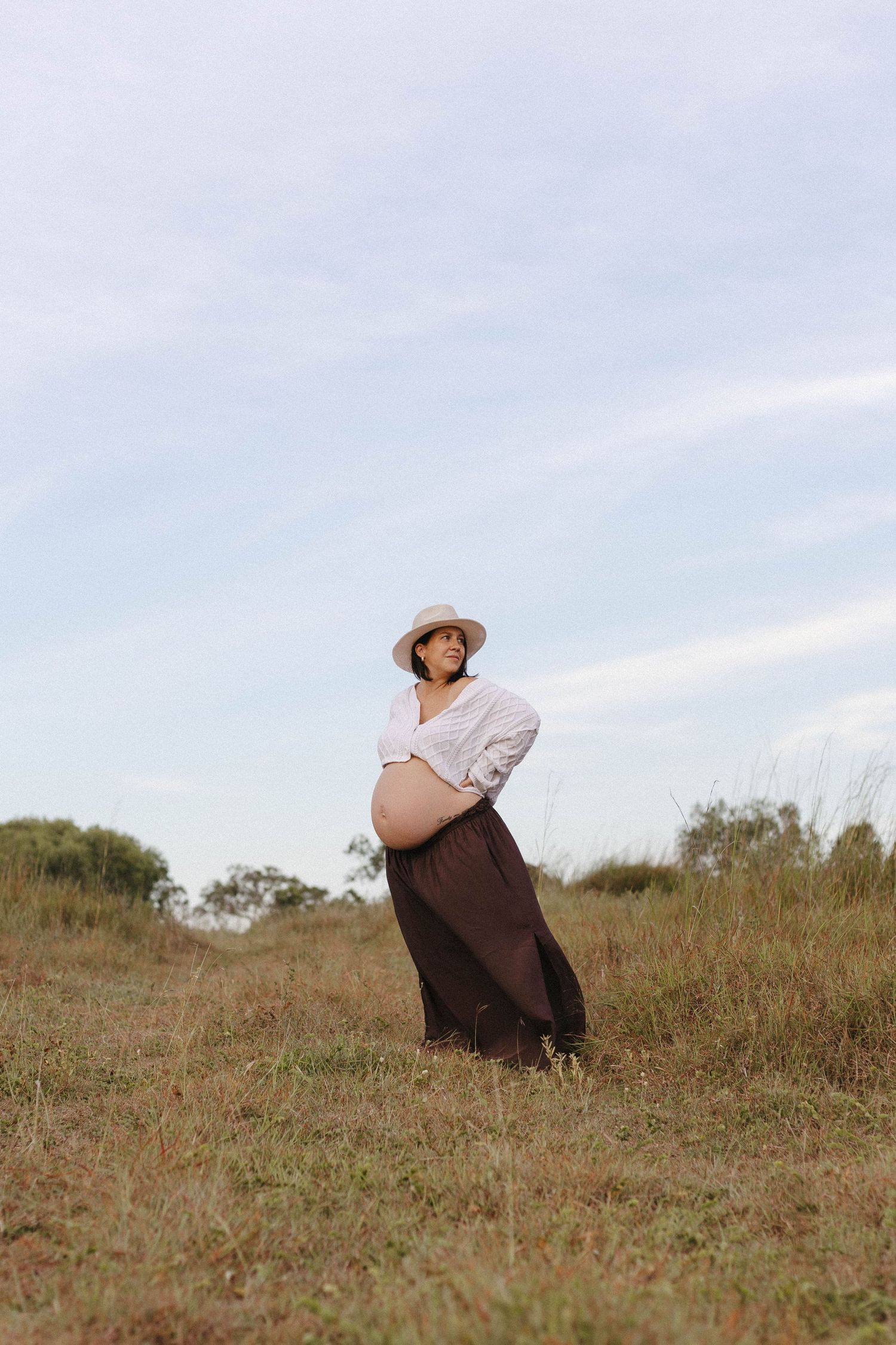 Figure in a brown dress and sun hat sits peacefully on a hillside overlooking natural grassland.