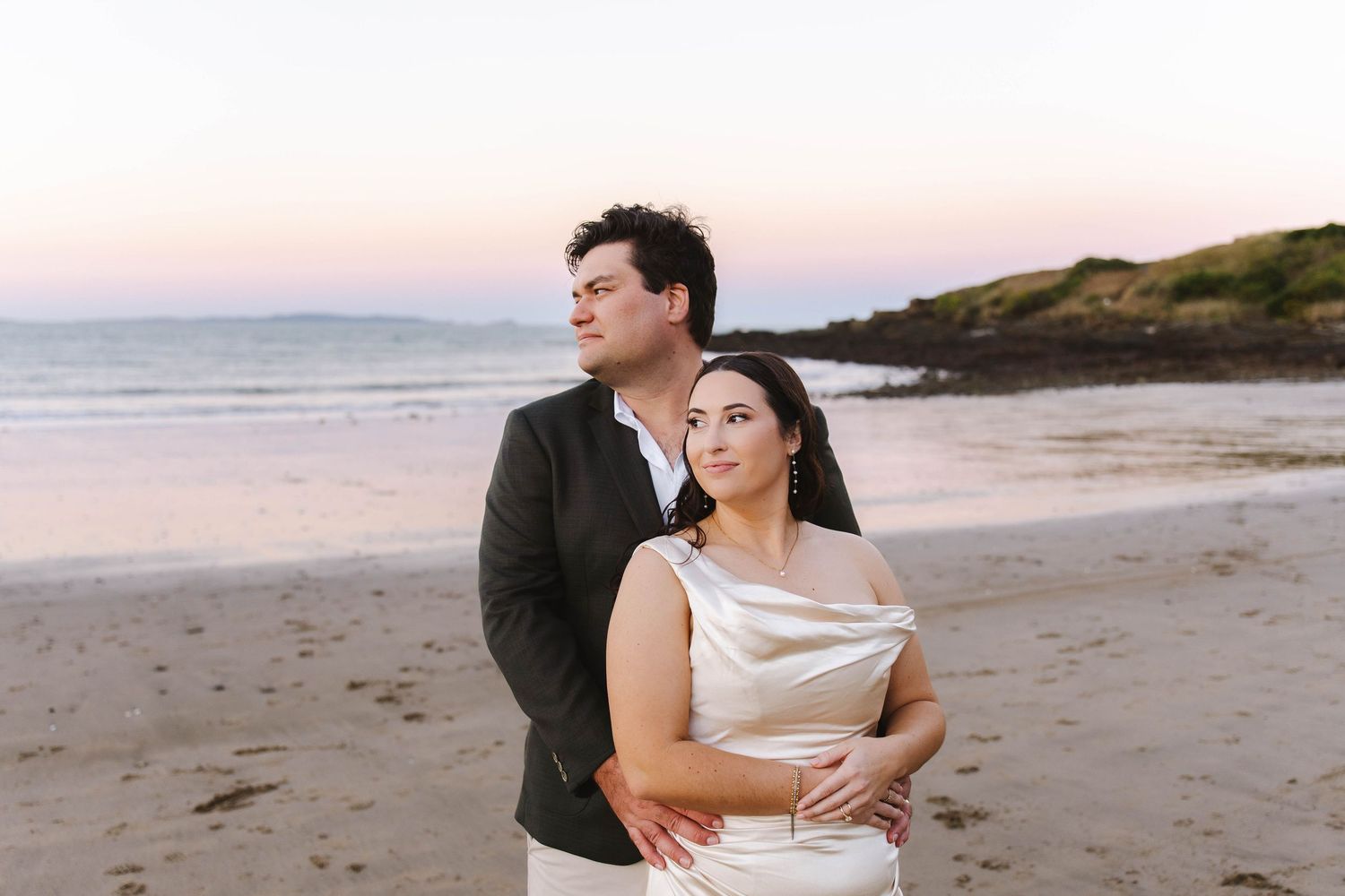 Couple embracing on a beach at sunset, woman in white dress, man in dark jacket, soft pink sky and ocean backdrop.
