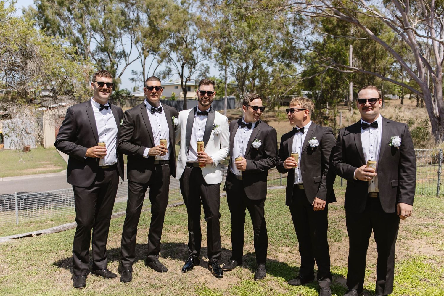 Wedding party in black tuxedos toasting with drinks outdoors on a sunny day.