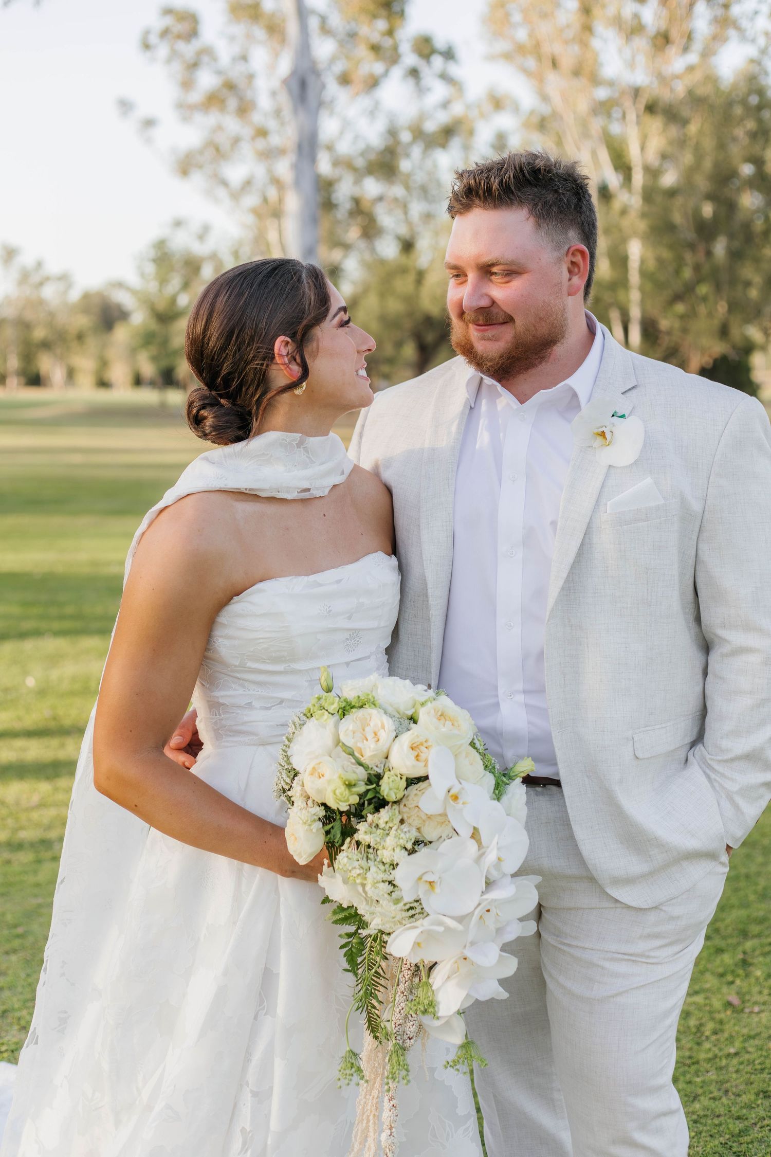 Elegant couple in all-white attire pose together on grassy field with white floral bouquet.