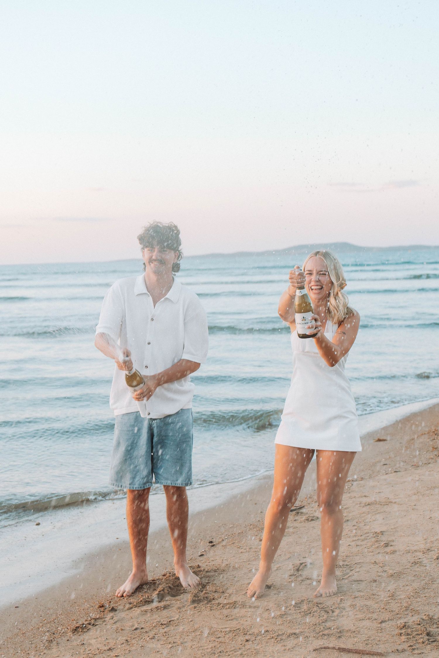 Couple celebrating on a beach at sunset, popping champagne bottles while wearing white outfits, with ocean waves behind them.