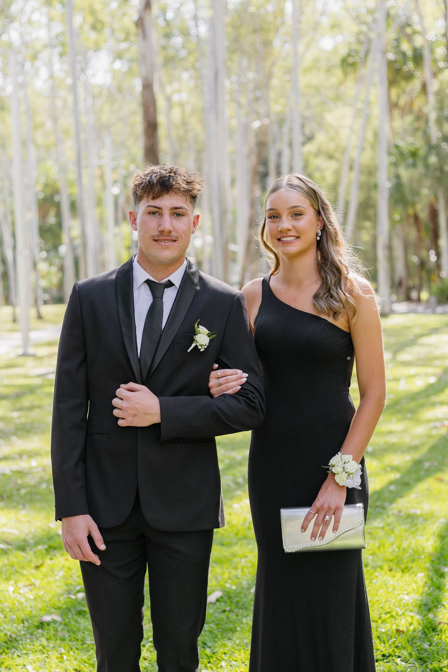 A couple in formal black attire poses together in a wooded outdoor setting with sunlit trees in the background.