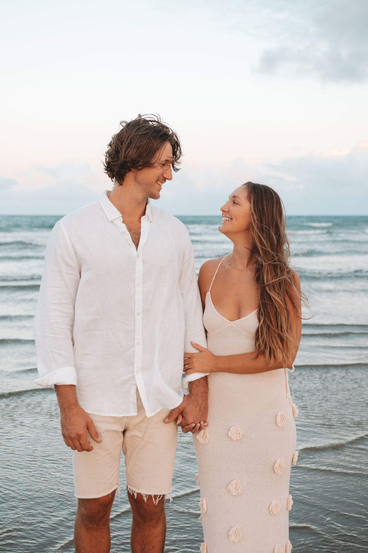 Couple in white outfits walking hand in hand along beach at sunset.