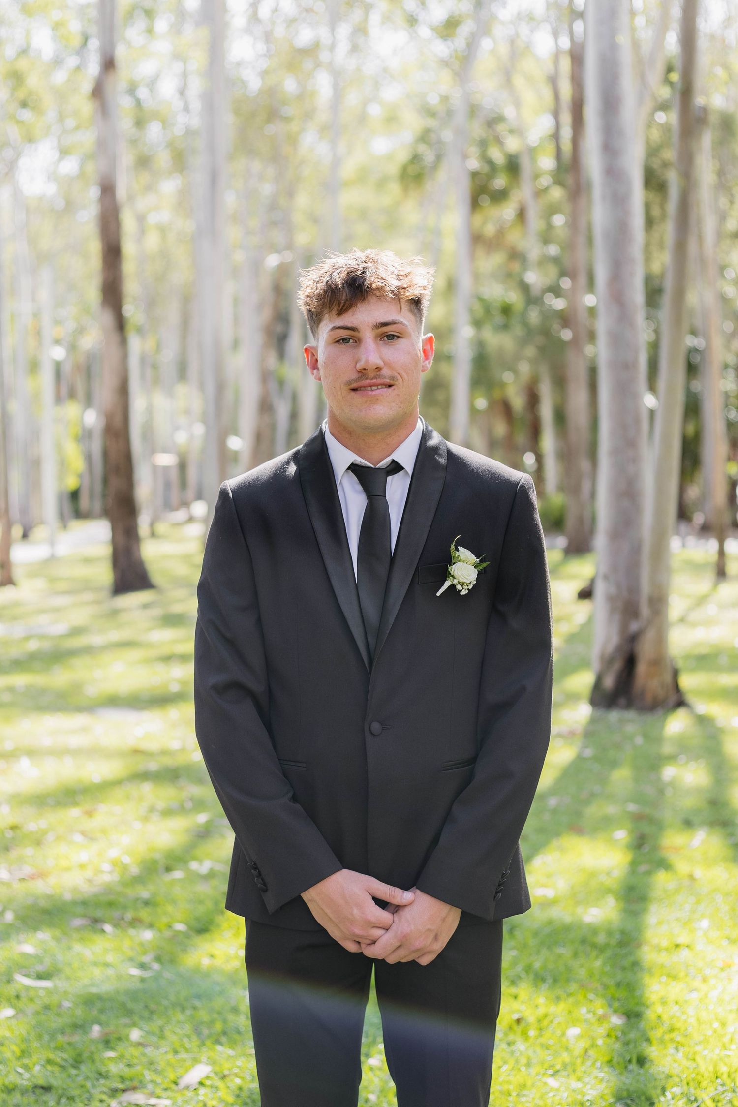 A person in a black suit and tie stands in a wooded area with sunlit trees in the background.