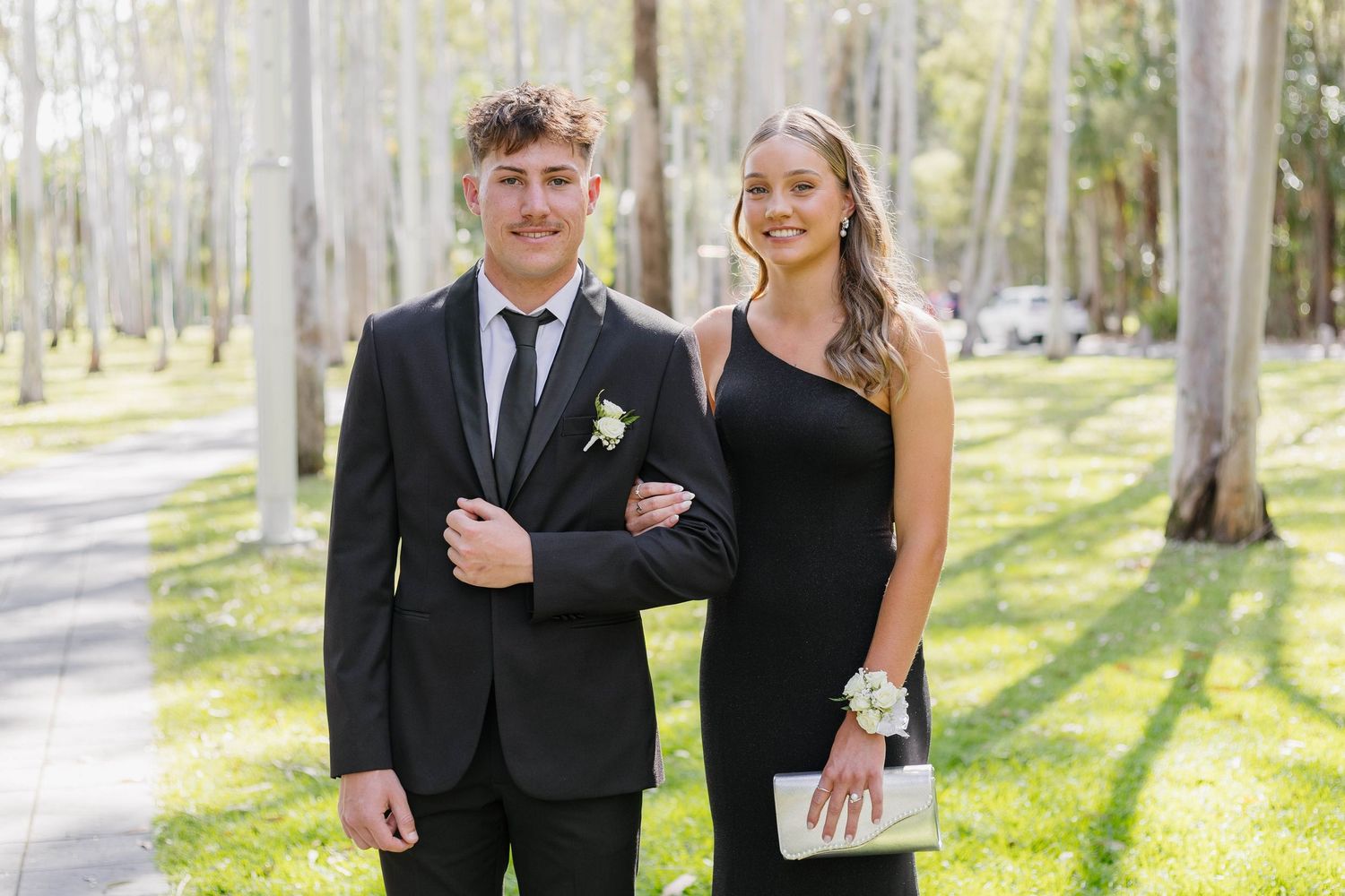 Elegant couple in formal attire poses together in a sunlit forest setting.