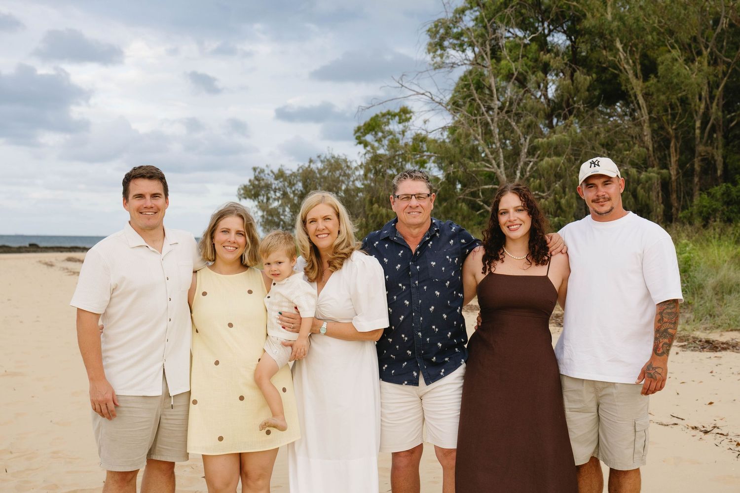 Extended family wearing coordinated white and neutral outfits poses on beach.