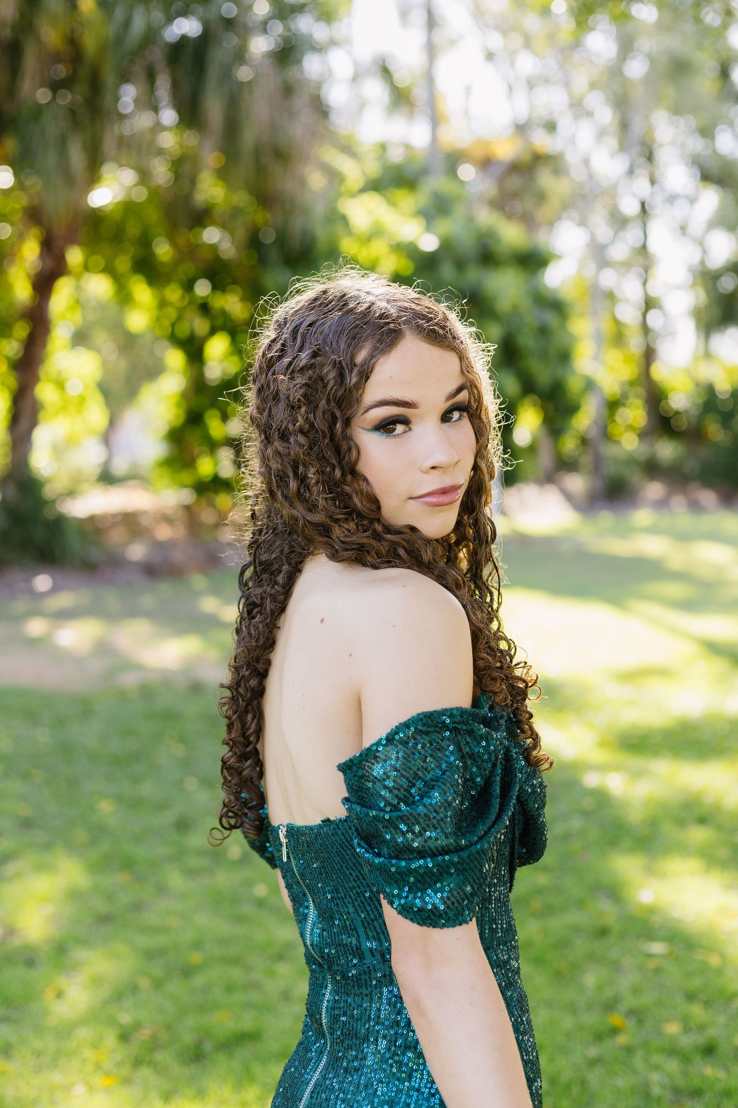 Woman in off-shoulder emerald sequin gown posing in garden setting.