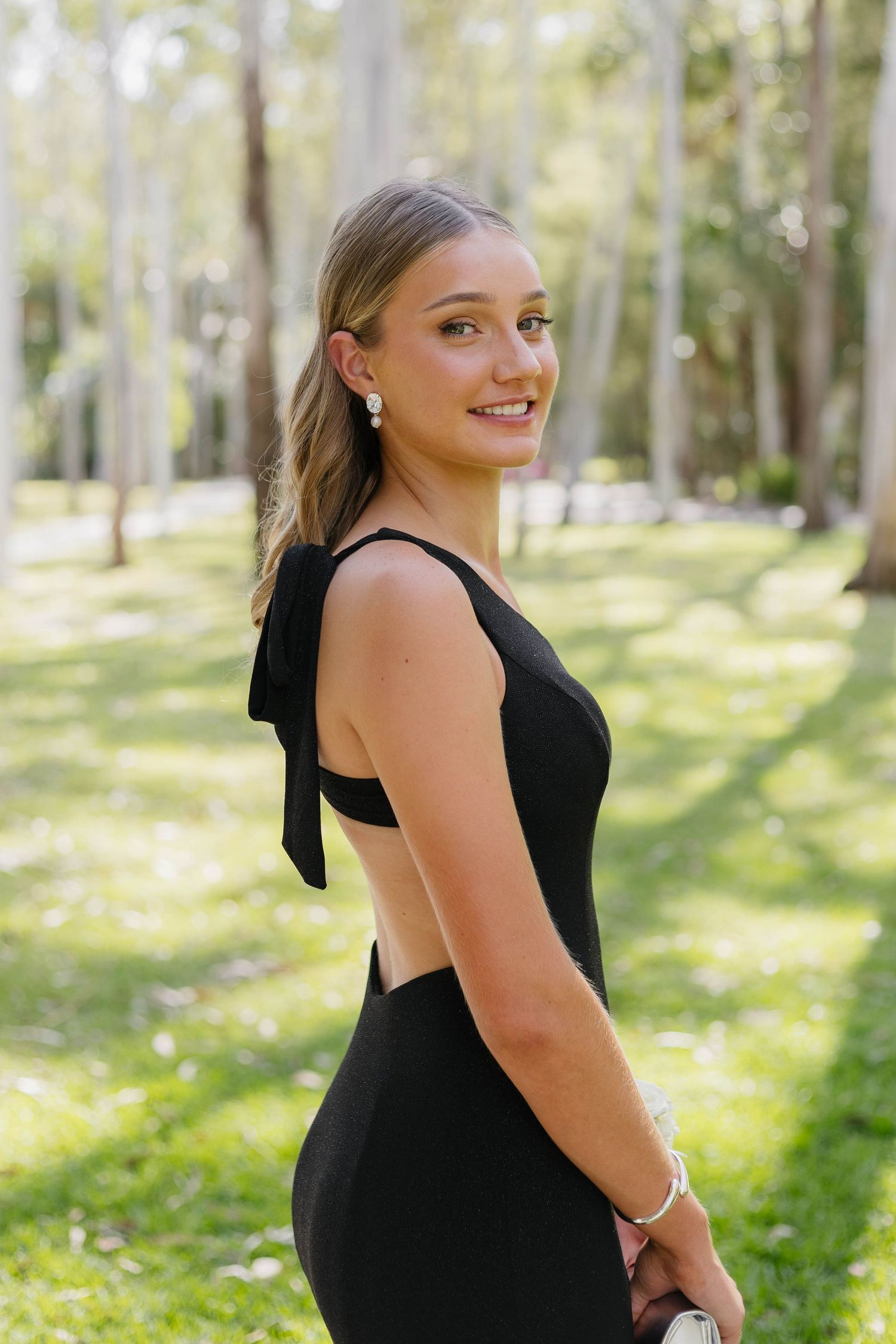 A person in a black formal dress poses outdoors in a wooded setting with natural lighting.