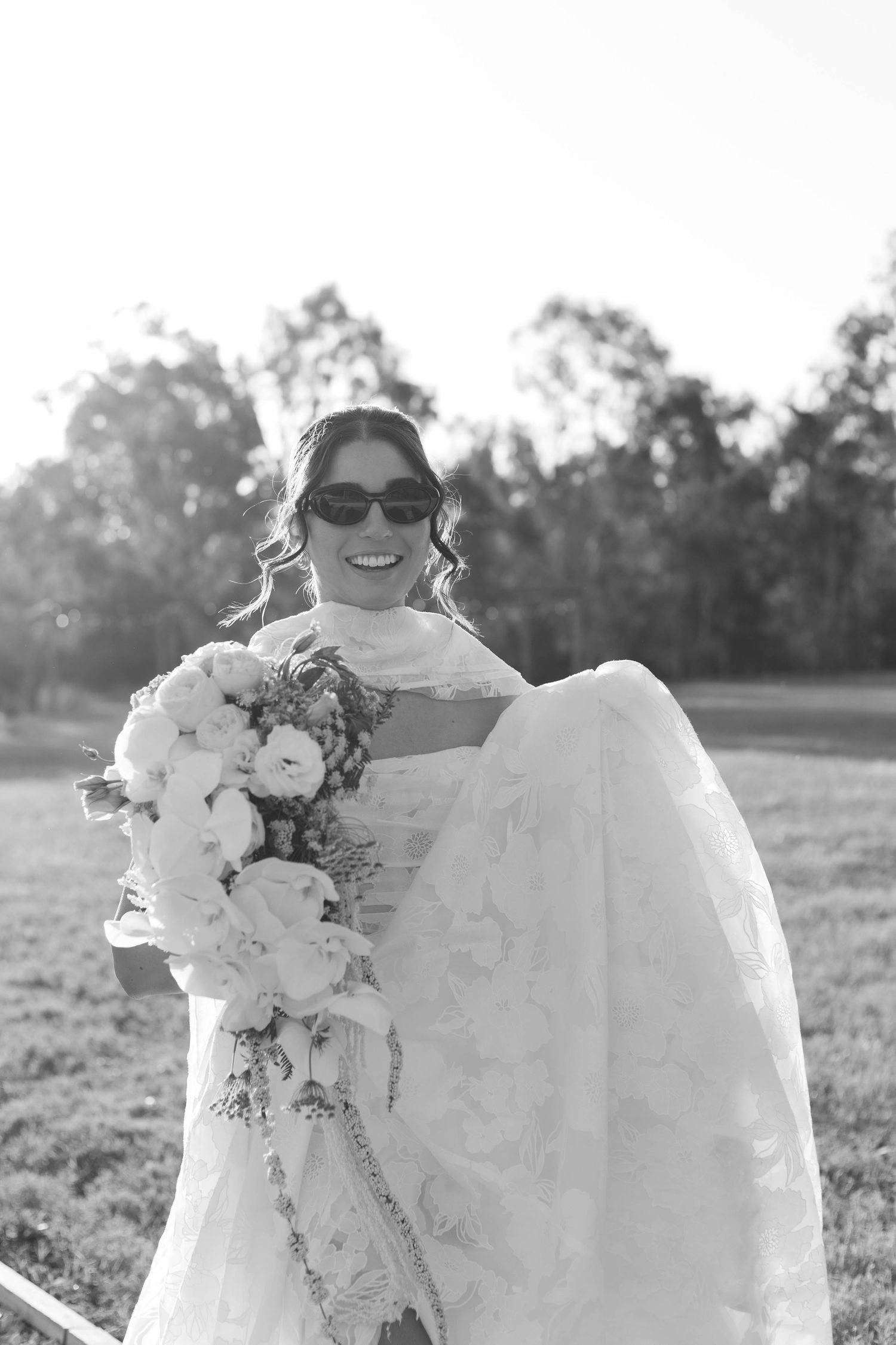 A joyful bride in sunglasses holds a cascading white bouquet during an outdoor wedding photoshoot.