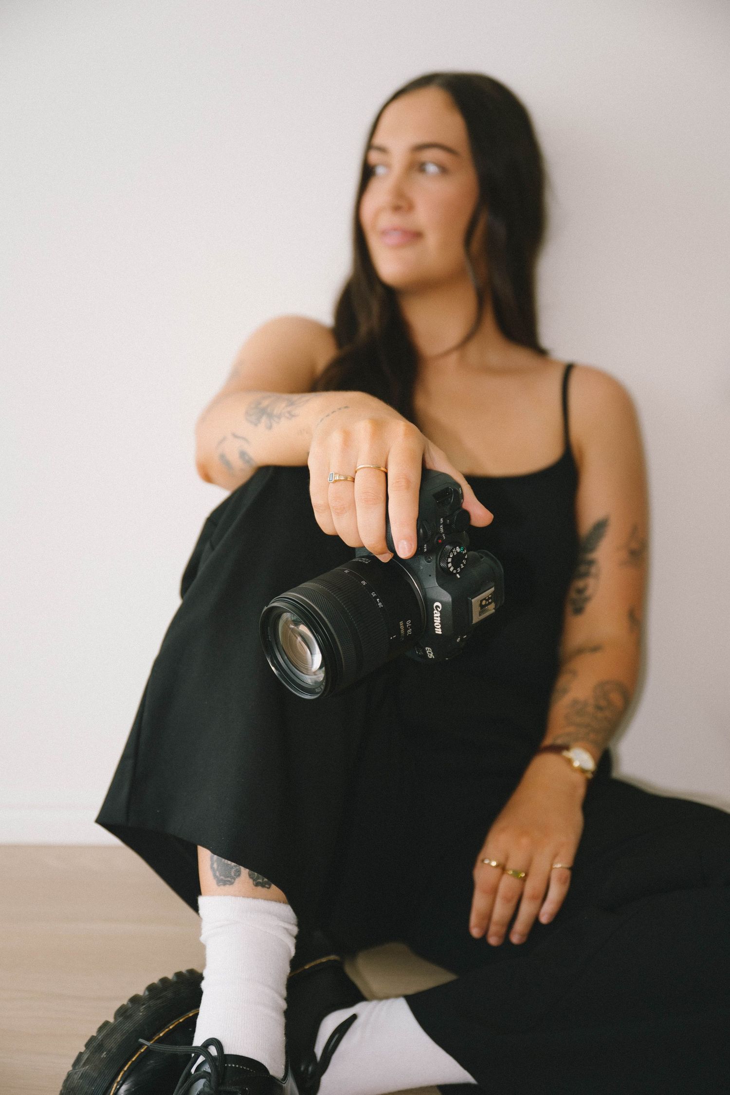 A person in black clothing sits against a white wall while holding a camera in their hands.