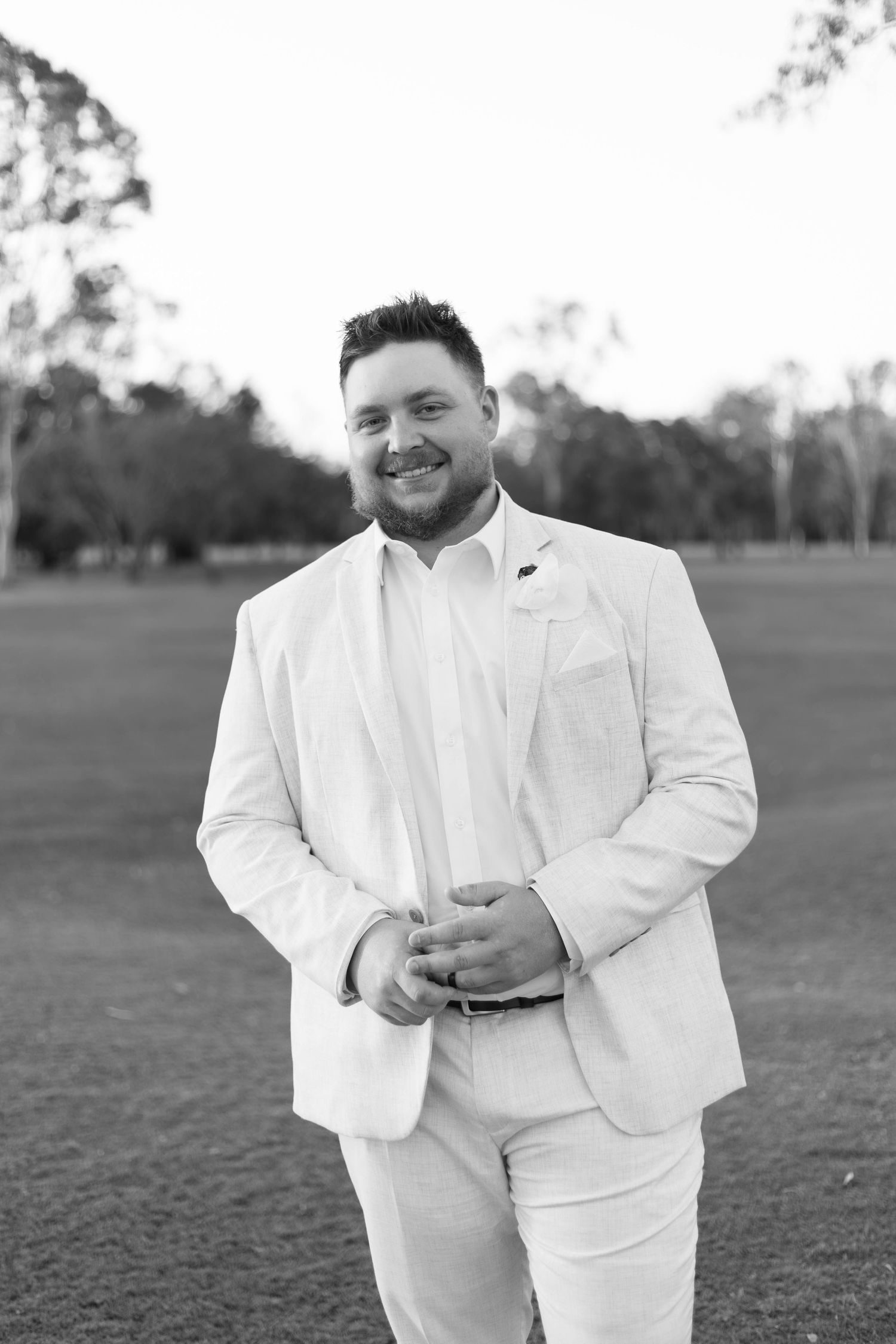 Groom in crisp white suit poses on grassy field in black and white photo.