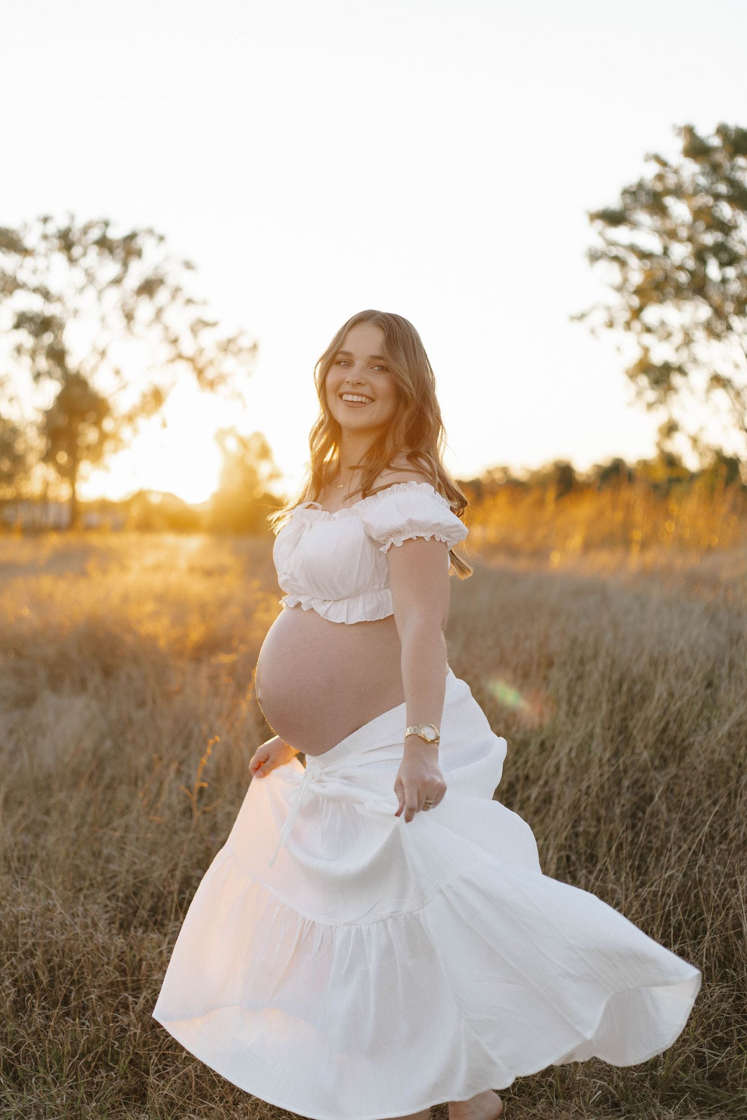 A flowing white dress twirls during a sunset maternity photoshoot in a field.
