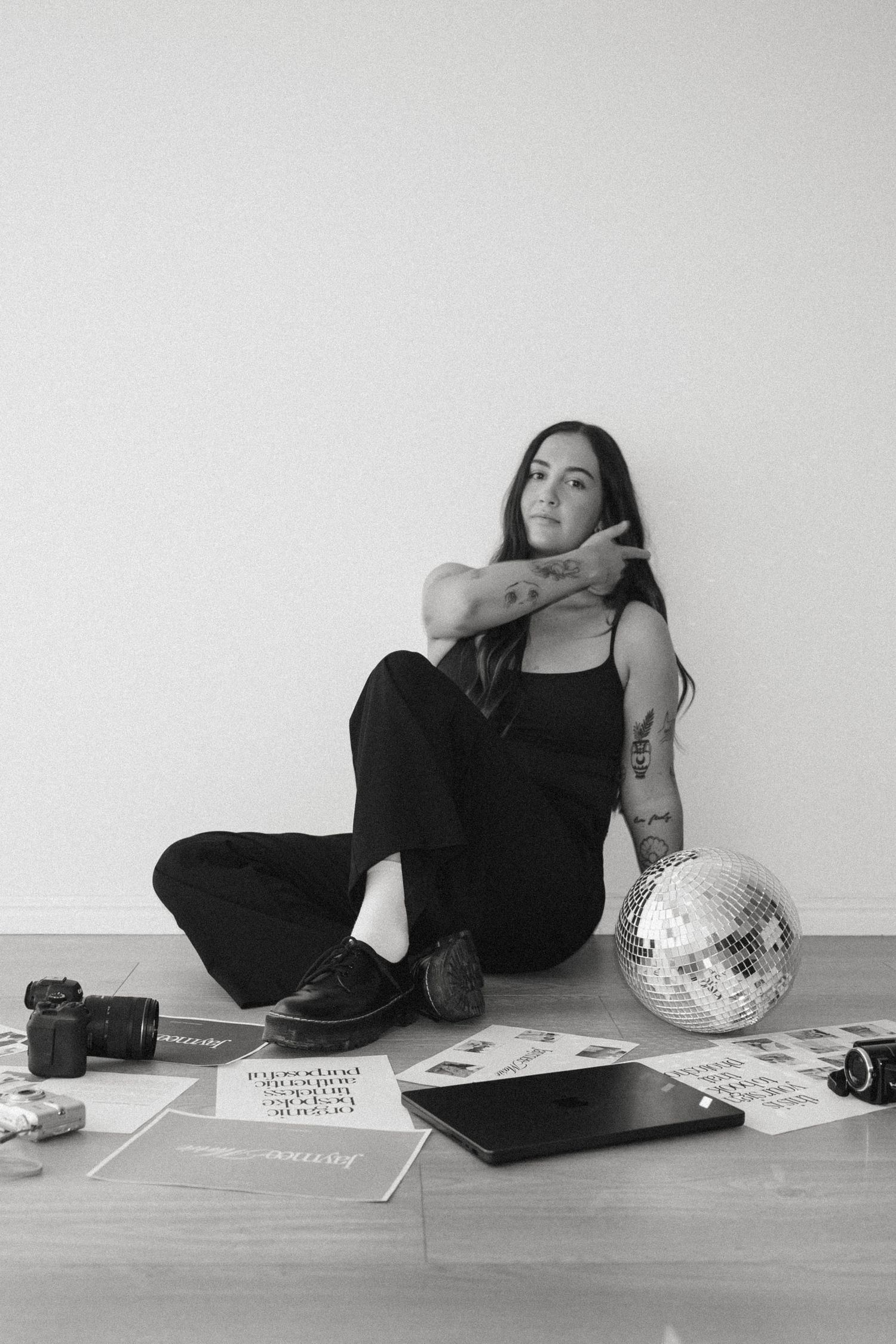 A person in black clothing lies on the floor beside a disco ball and other objects in a minimalist setting.