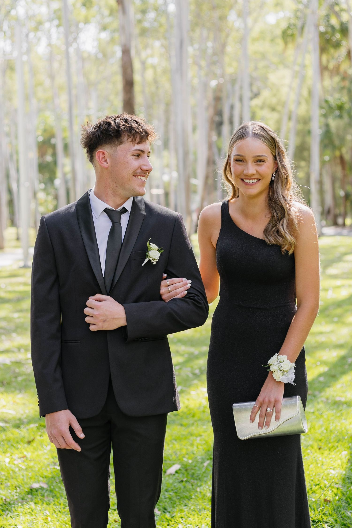 Formal couple in black attire poses in sunny woodland setting.