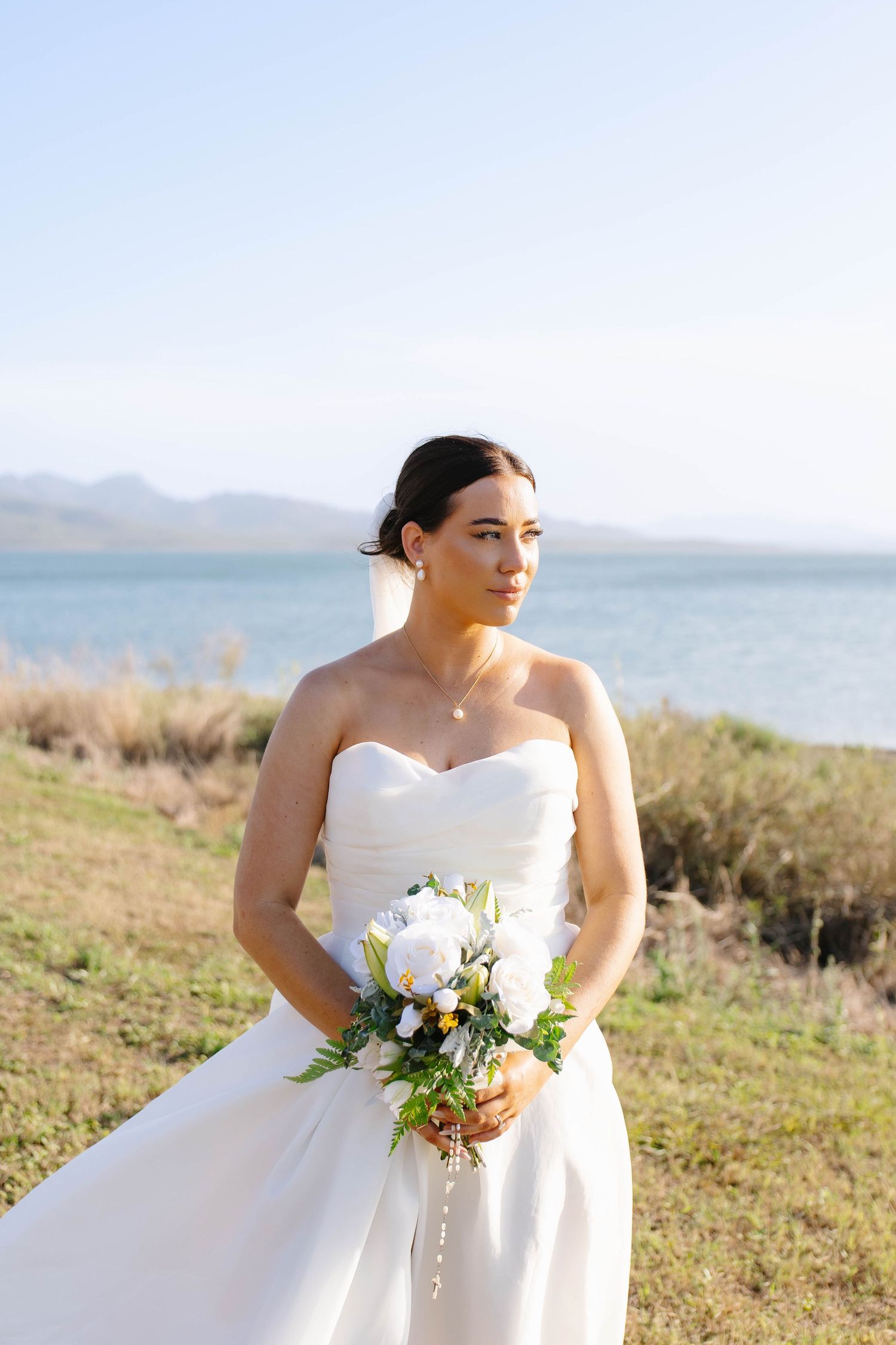 A bride in a white strapless gown holds a delicate bouquet against a waterfront landscape.