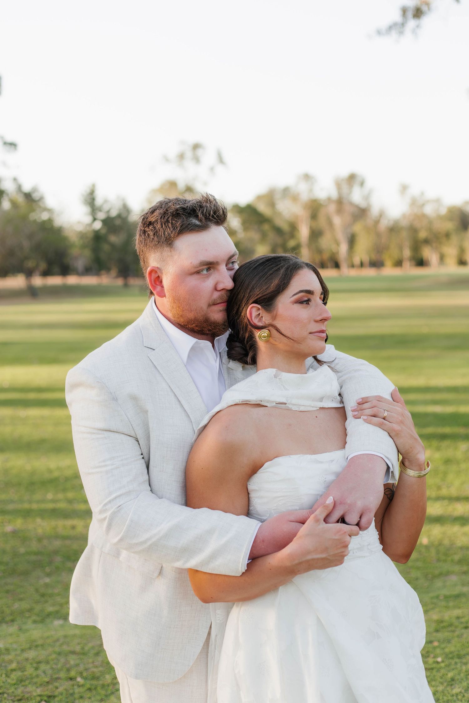 A romantic wedding couple embraces in white attire on a sunlit grassy field.