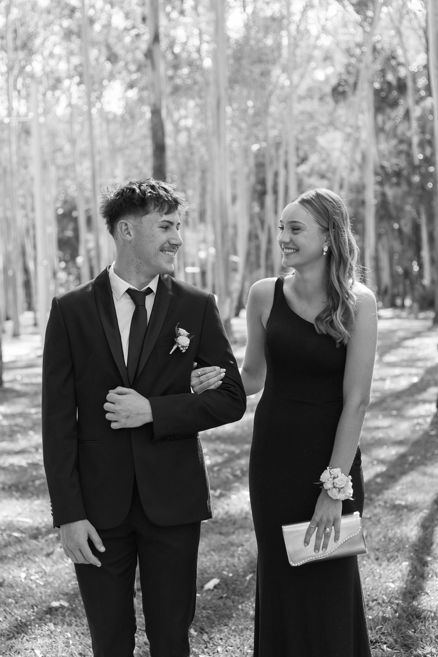 A couple shares a candid moment laughing together while dressed formally in a sunlit forest setting.