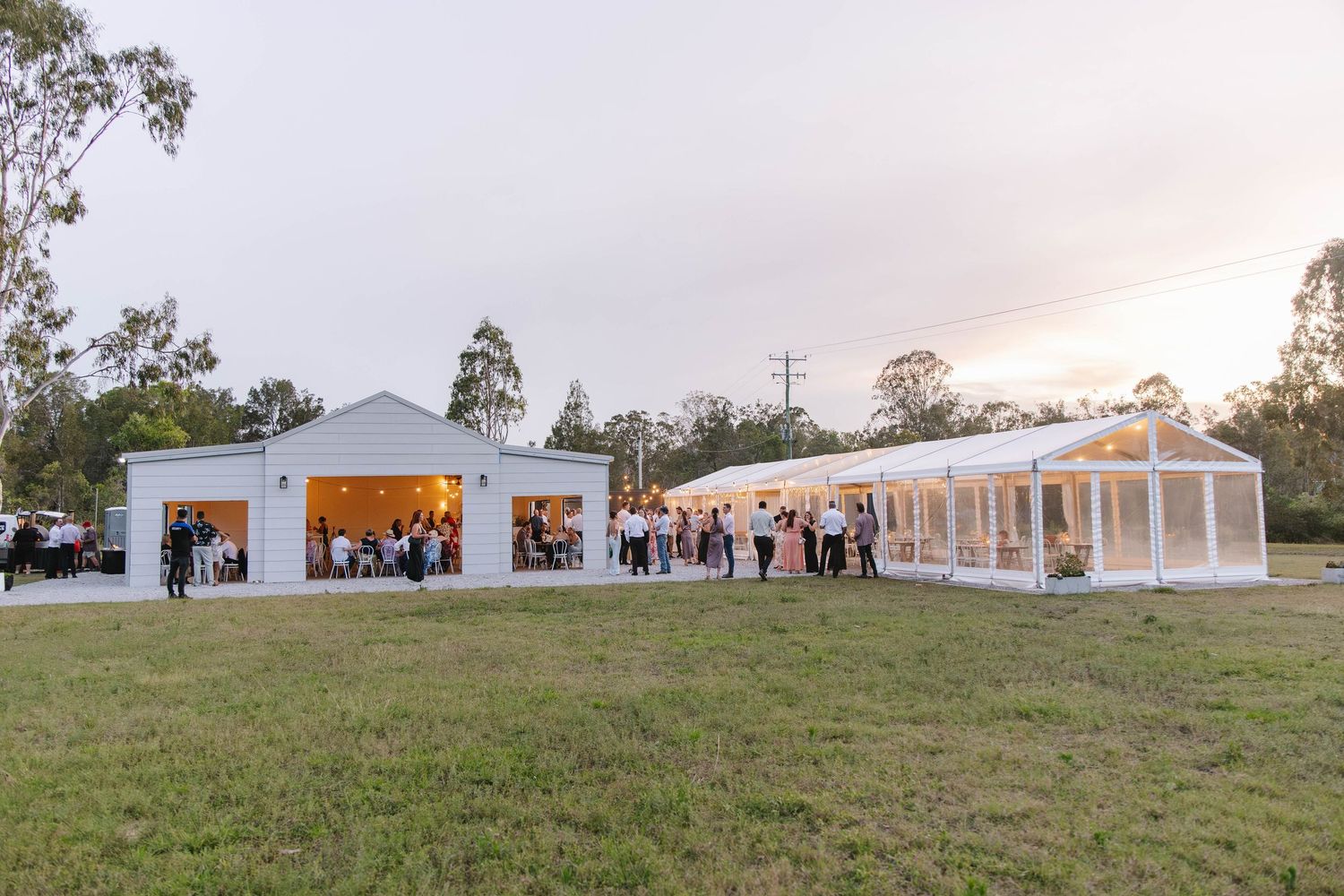 An elegant white wedding venue glows at dusk with guests mingling outside.