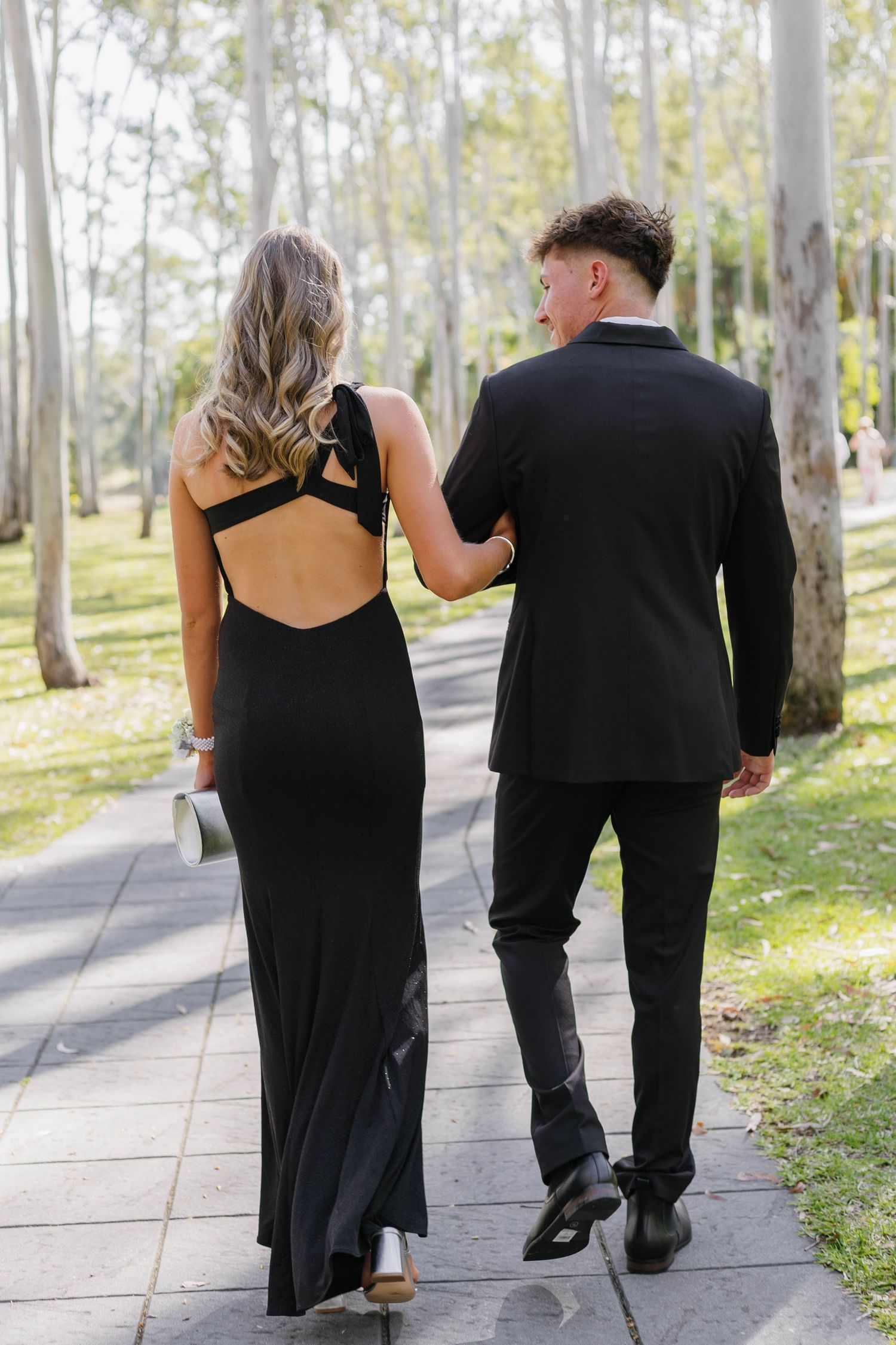 A couple walking together on a path through a sunlit wooded area wearing formal black attire.