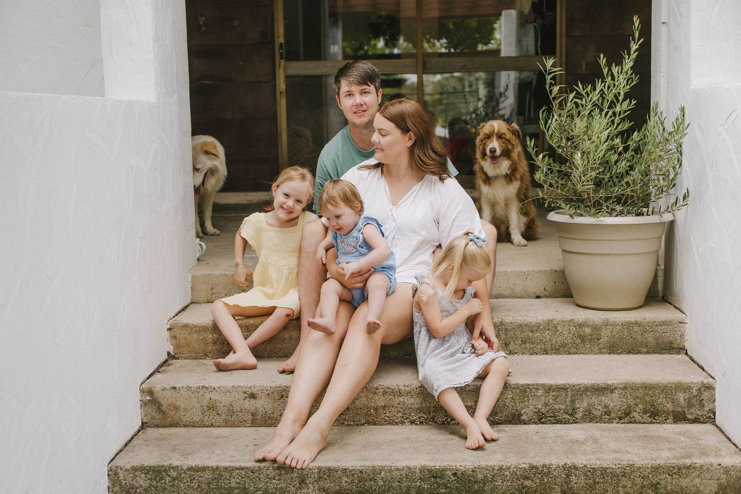 Family sitting casually on front porch steps of a house with potted plants.