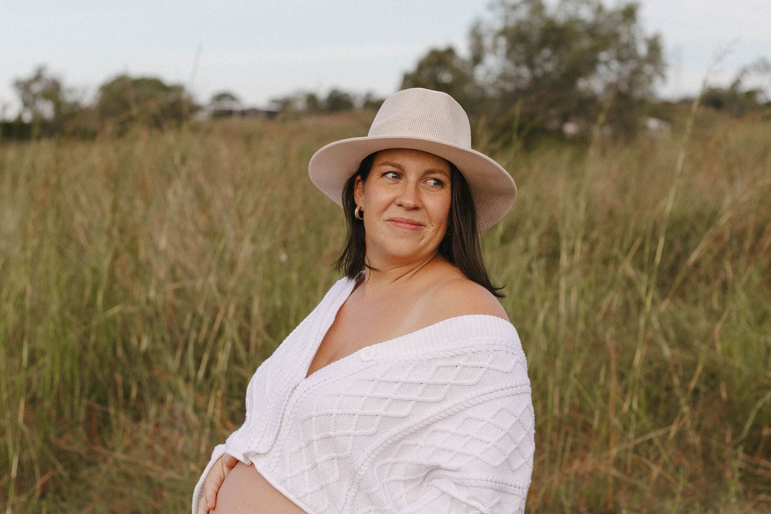 A person in a wide-brimmed hat and white dress poses thoughtfully in a grassy field during golden hour.