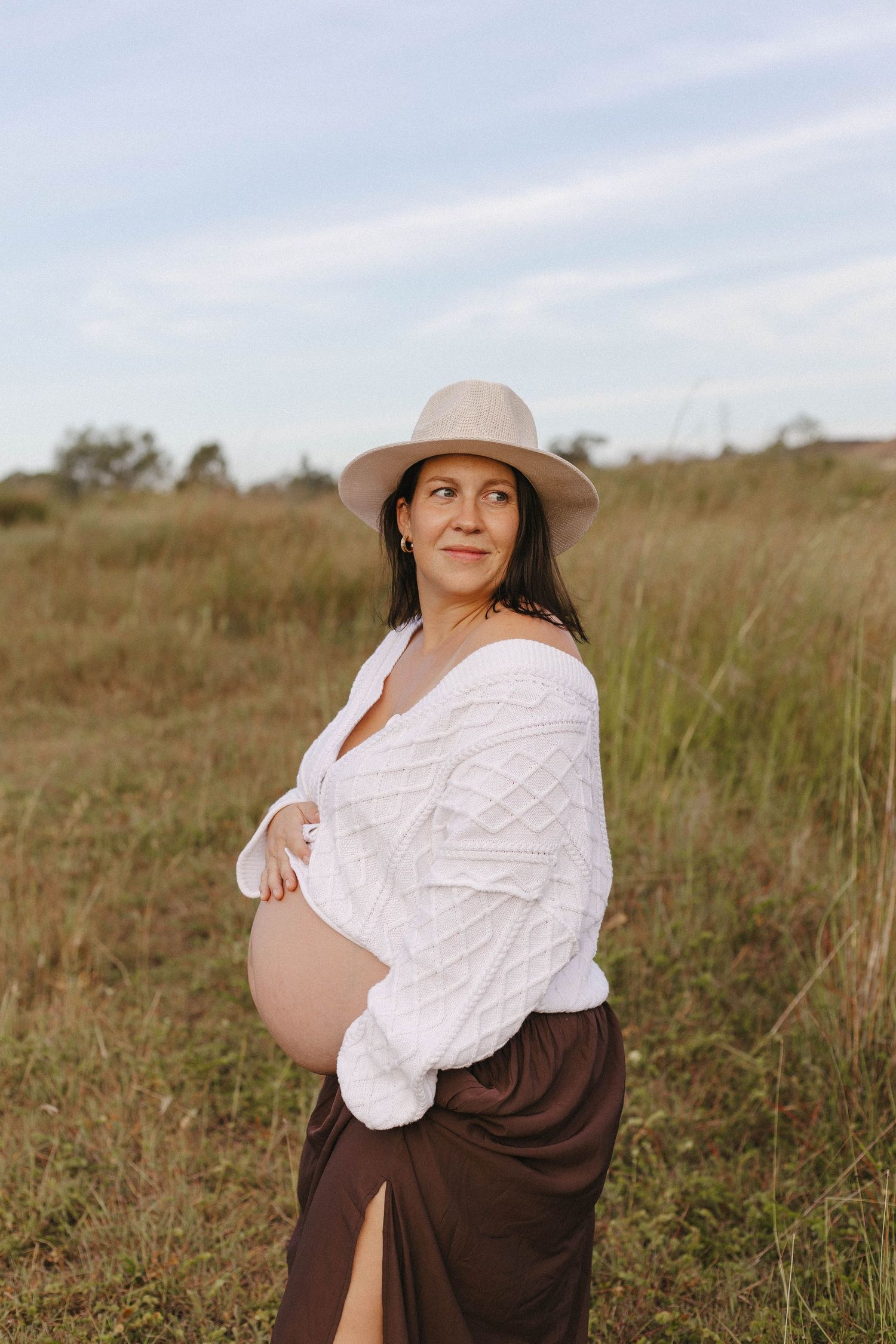 A person in a brown dress and beige hat poses thoughtfully in a grassy field during golden hour.