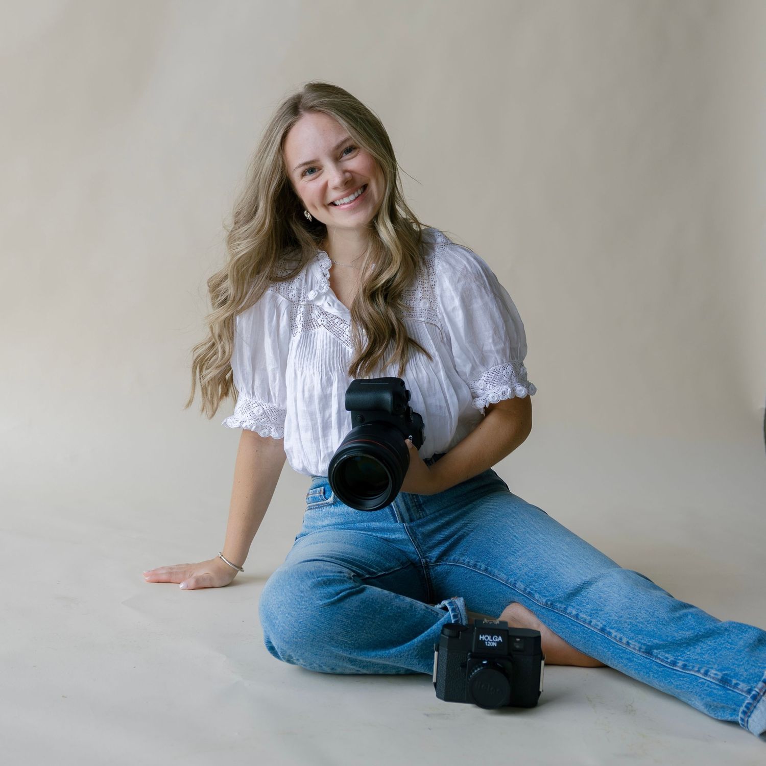 Smiling woman with long blonde hair sits on floor holding camera, wearing white blouse and jeans in bright studio.