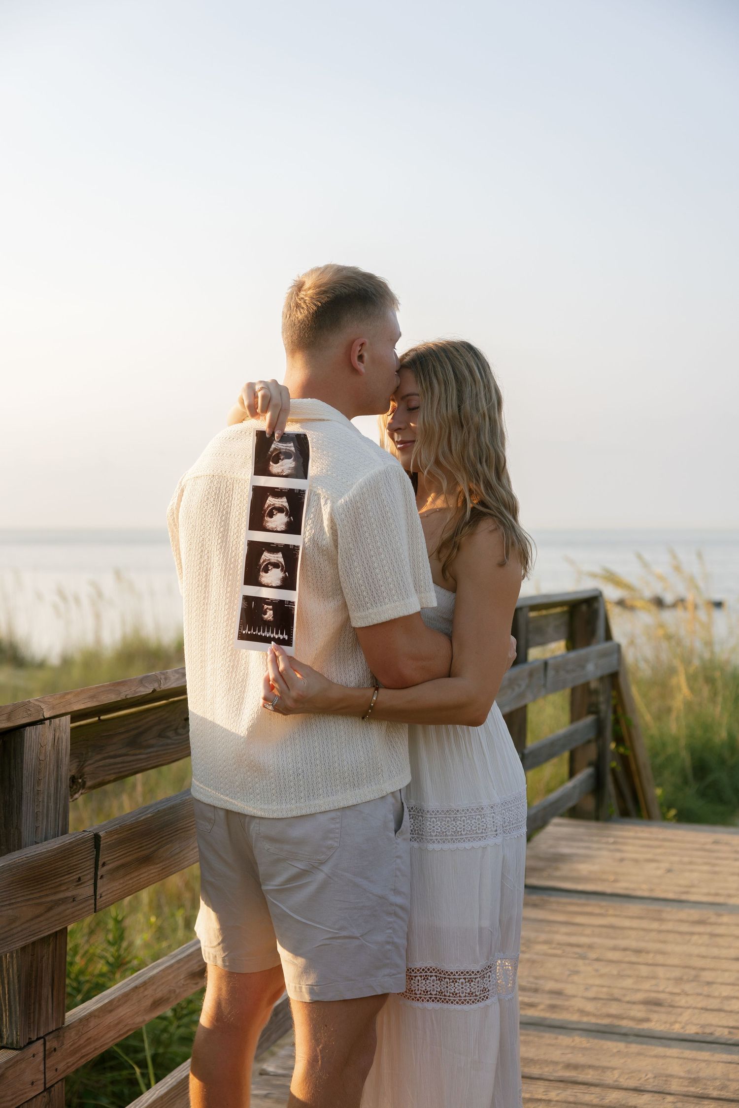 Couple embracing on a Norfolk Virginia beach boardwalk at sunset, holding ultrasound photos, announcing their pregnancy.