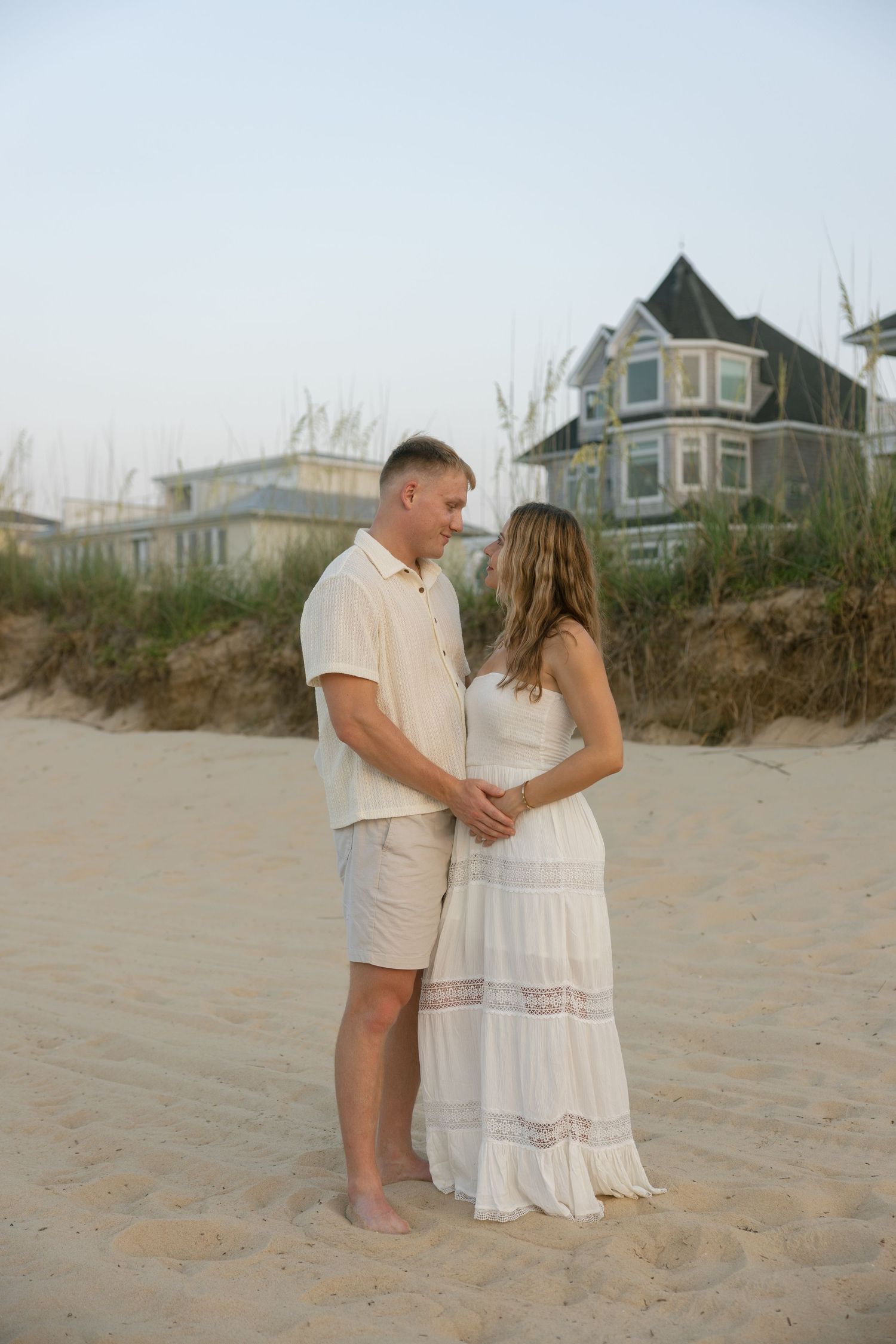 Couple in matching cream outfits standing on sandy beach with coastal houses and sea oats in background. Norfolk Virginia