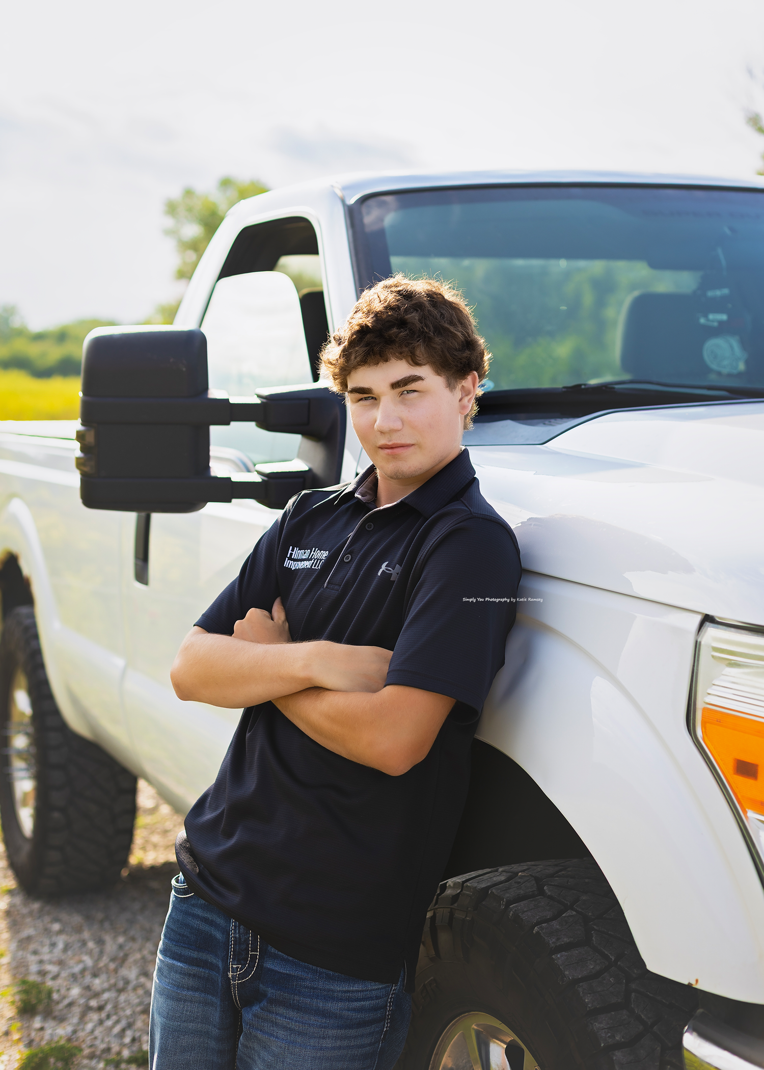 Young man in navy polo leaning against white pickup truck with arms crossed outdoors.
