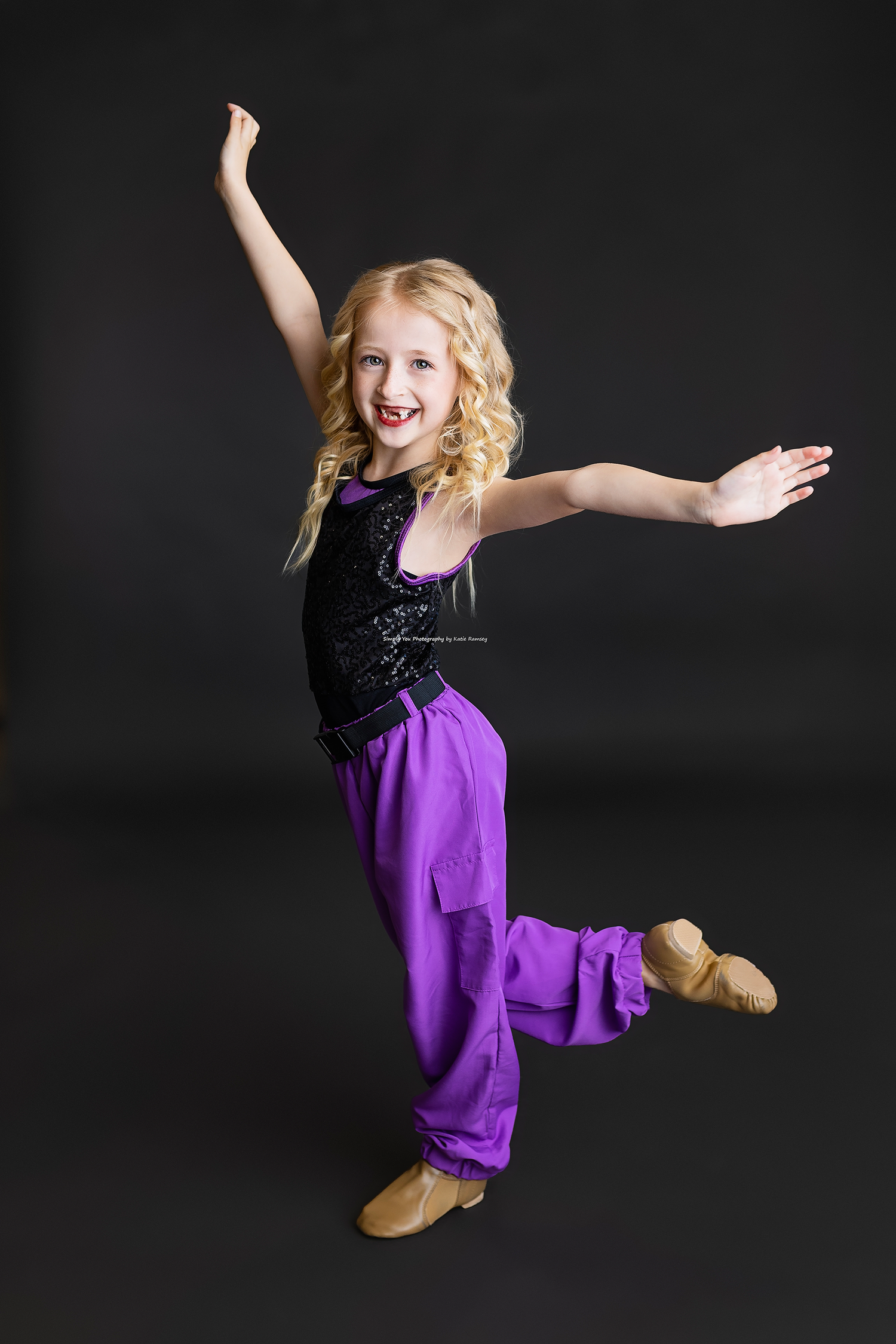 A young blonde girl in purple pants and black top strikes a joyful dance pose against a dark background.