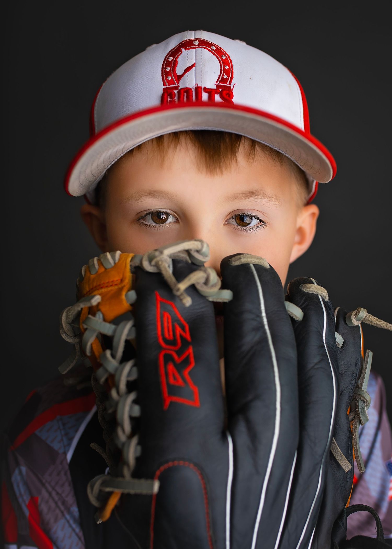 Young baseball player in Colts cap hiding face behind Rawlings baseball glove on dark background.