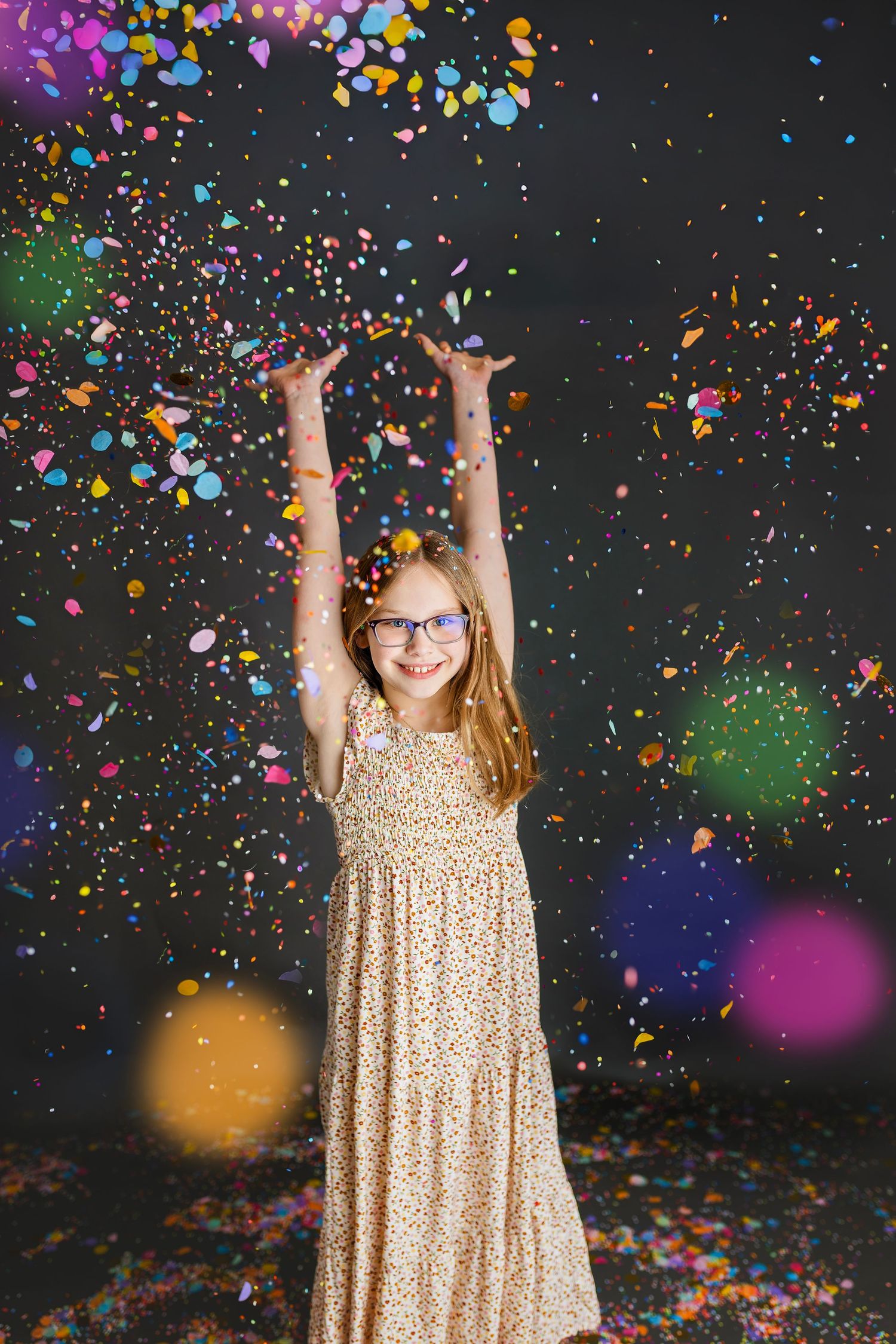 Happy girl in floral dress tossing colorful confetti in the air against a dark studio background.