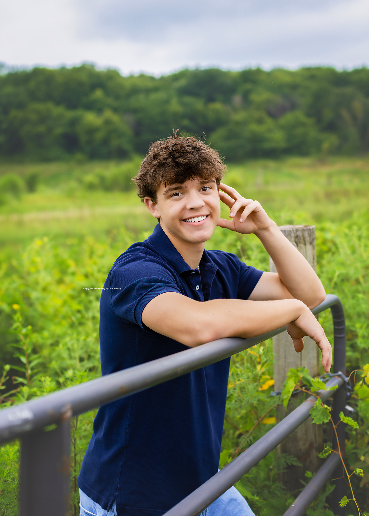 Smiling teenage boy in navy polo shirt leaning on metal railing with lush green field backdrop.