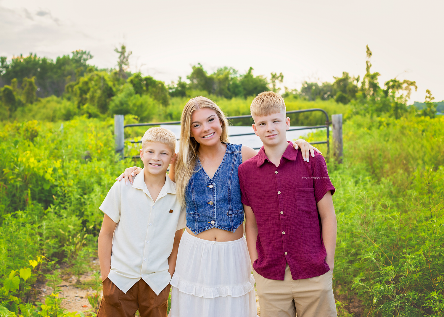 A woman in a denim vest and white skirt poses outdoors with two boys in a lush green field.