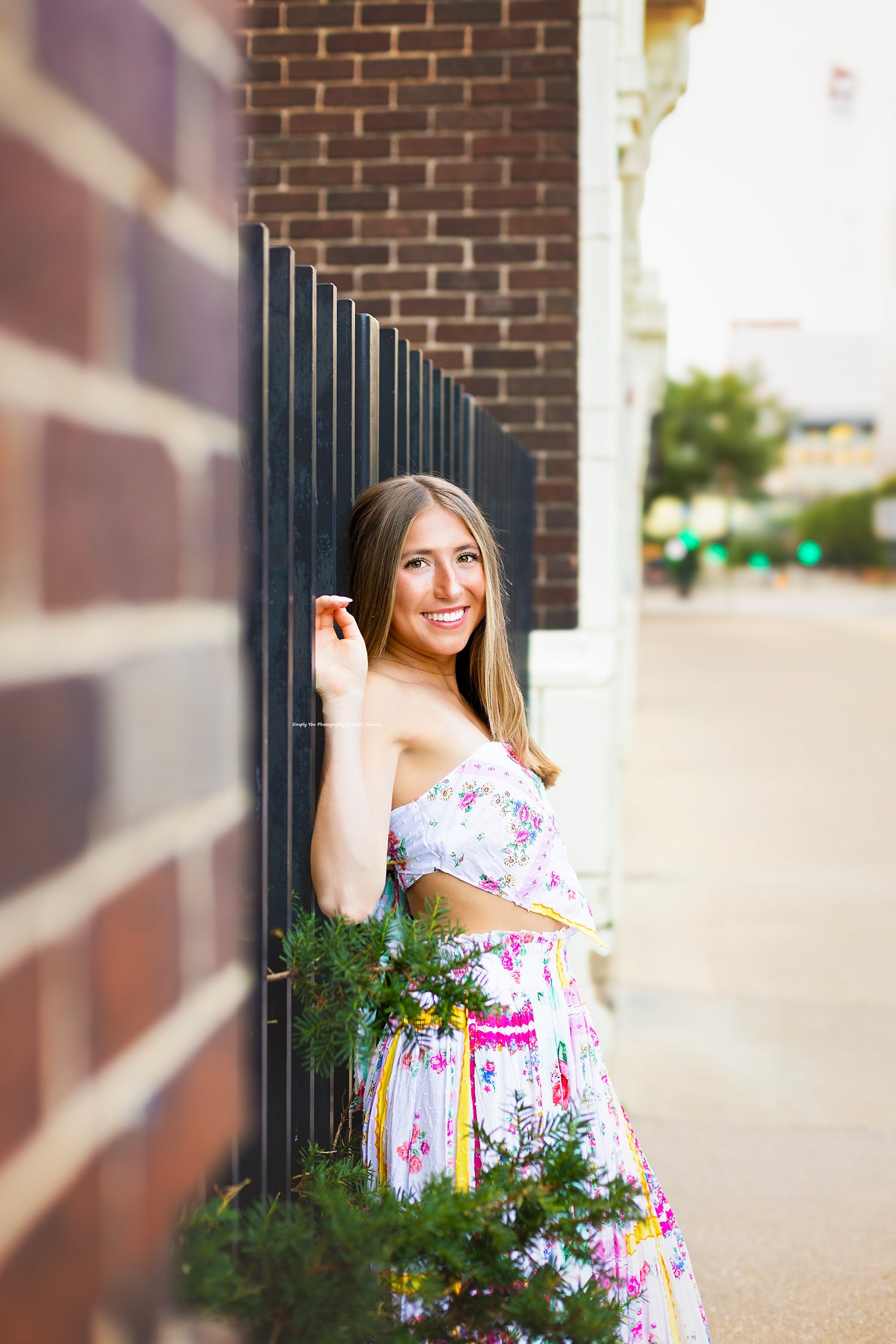 Smiling young woman in floral strapless dress posing beside brick wall and iron fence with greenery.