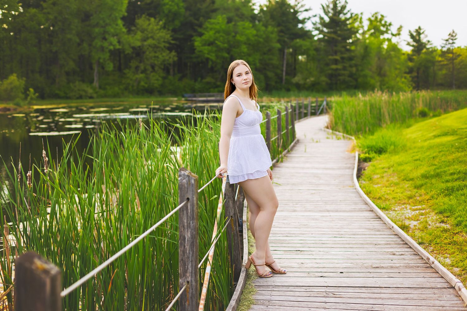 Young woman in white dress standing on a wooden boardwalk beside a serene pond with tall green reeds.