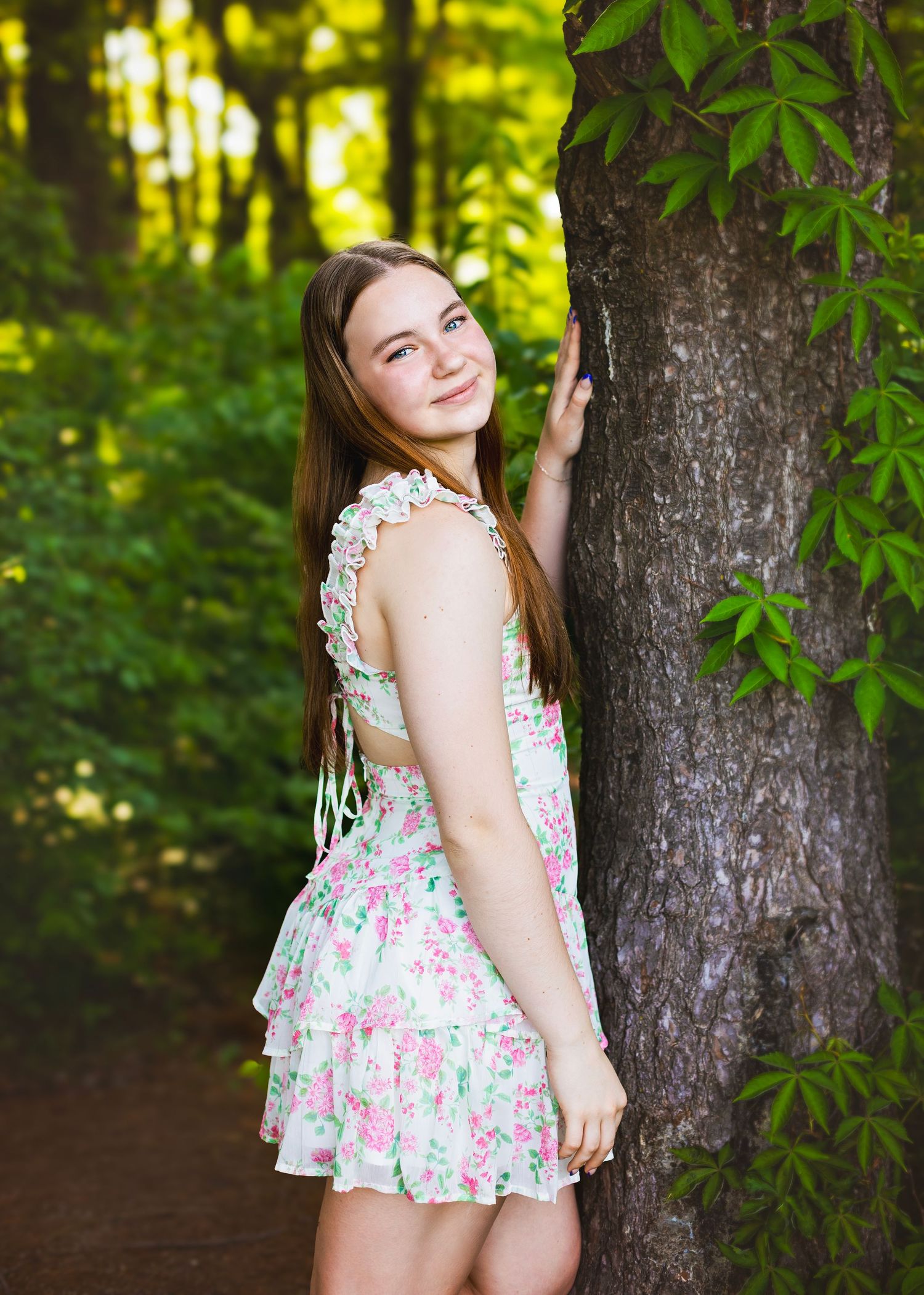 Young woman in floral mini dress smiling beside a tree in a lush green forest.