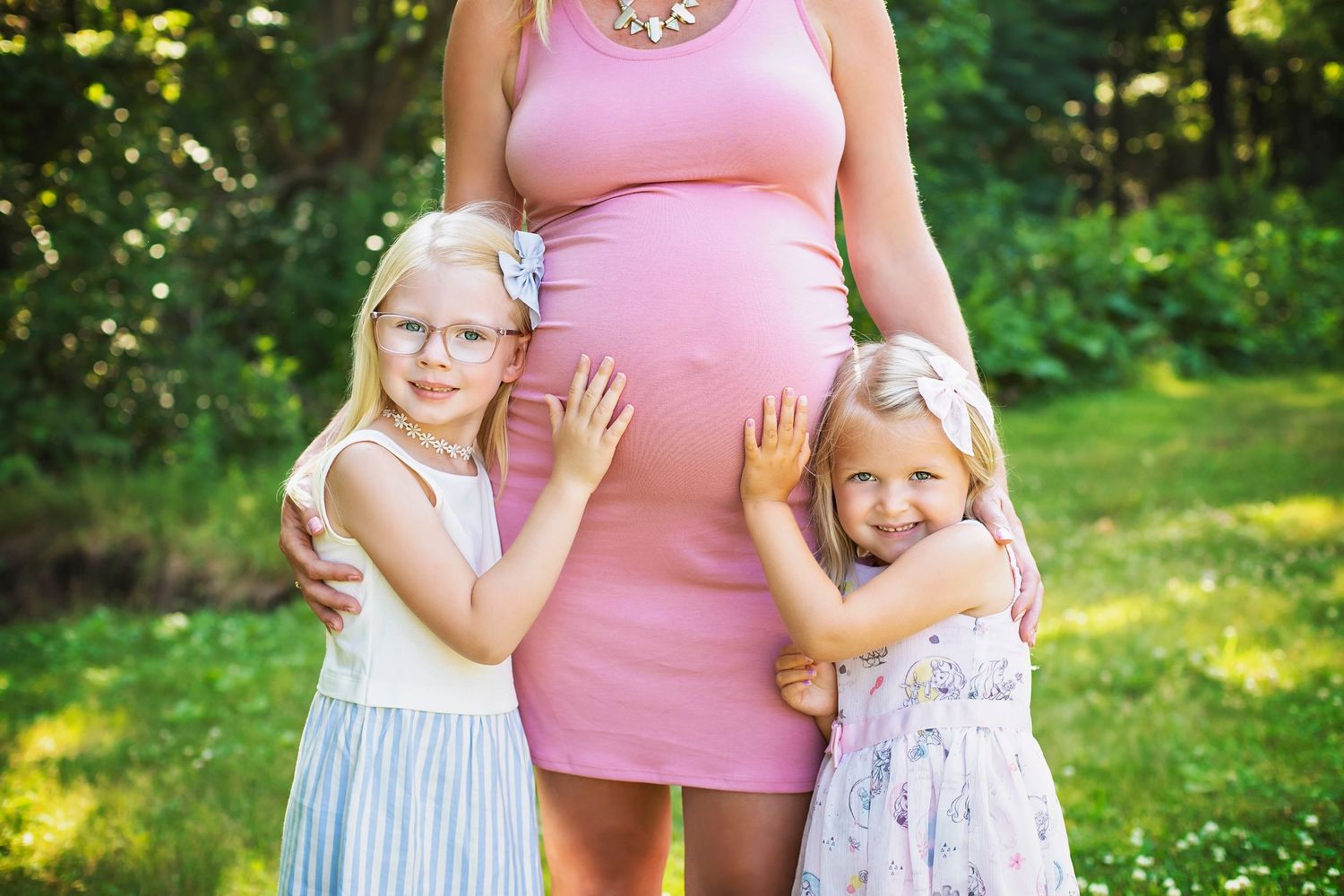 Pregnant woman in pink dress with two young blonde girls hugging her baby bump outdoors.