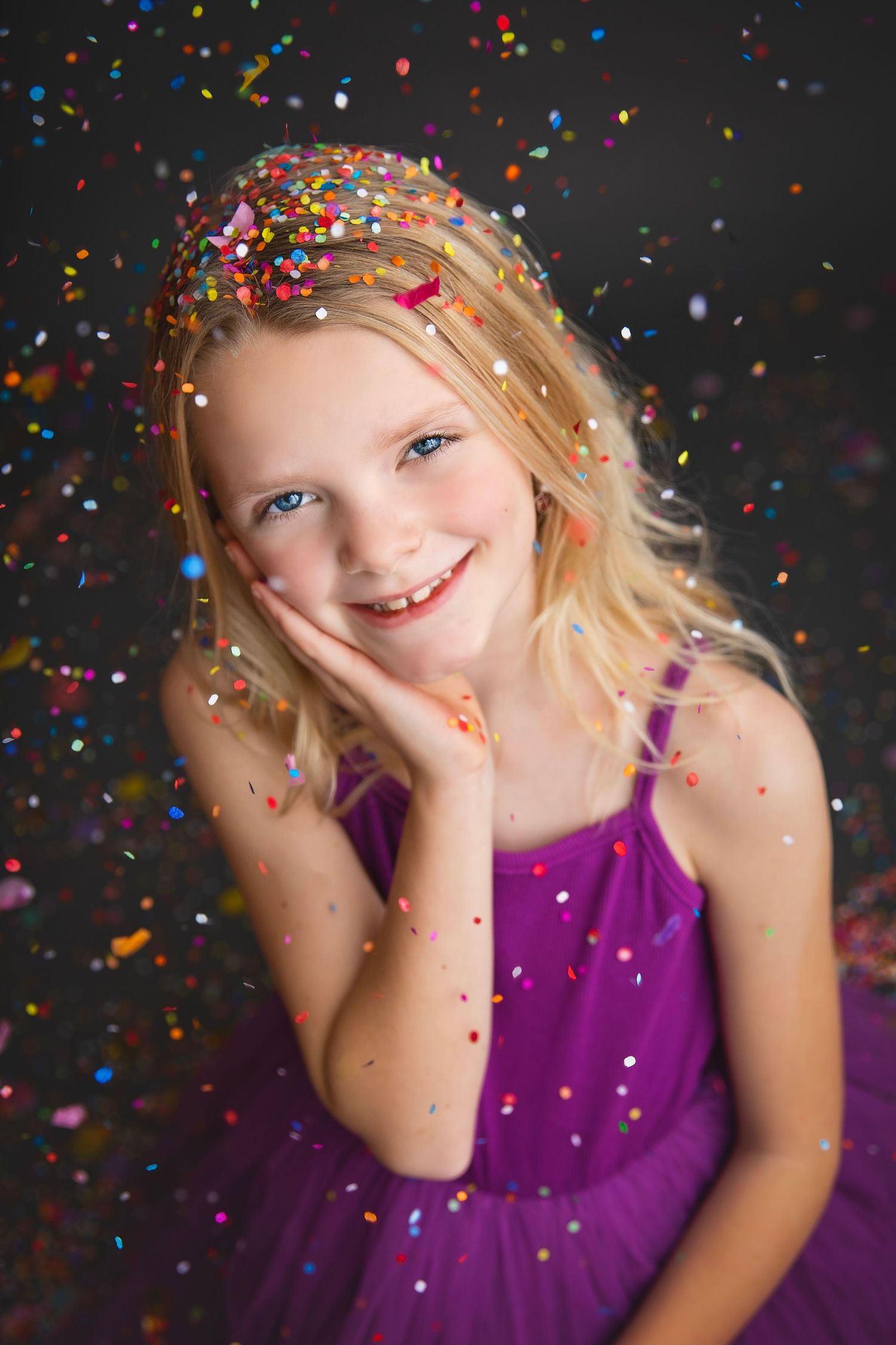 Smiling blonde girl in purple dress with colorful confetti falling around her in a studio portrait.