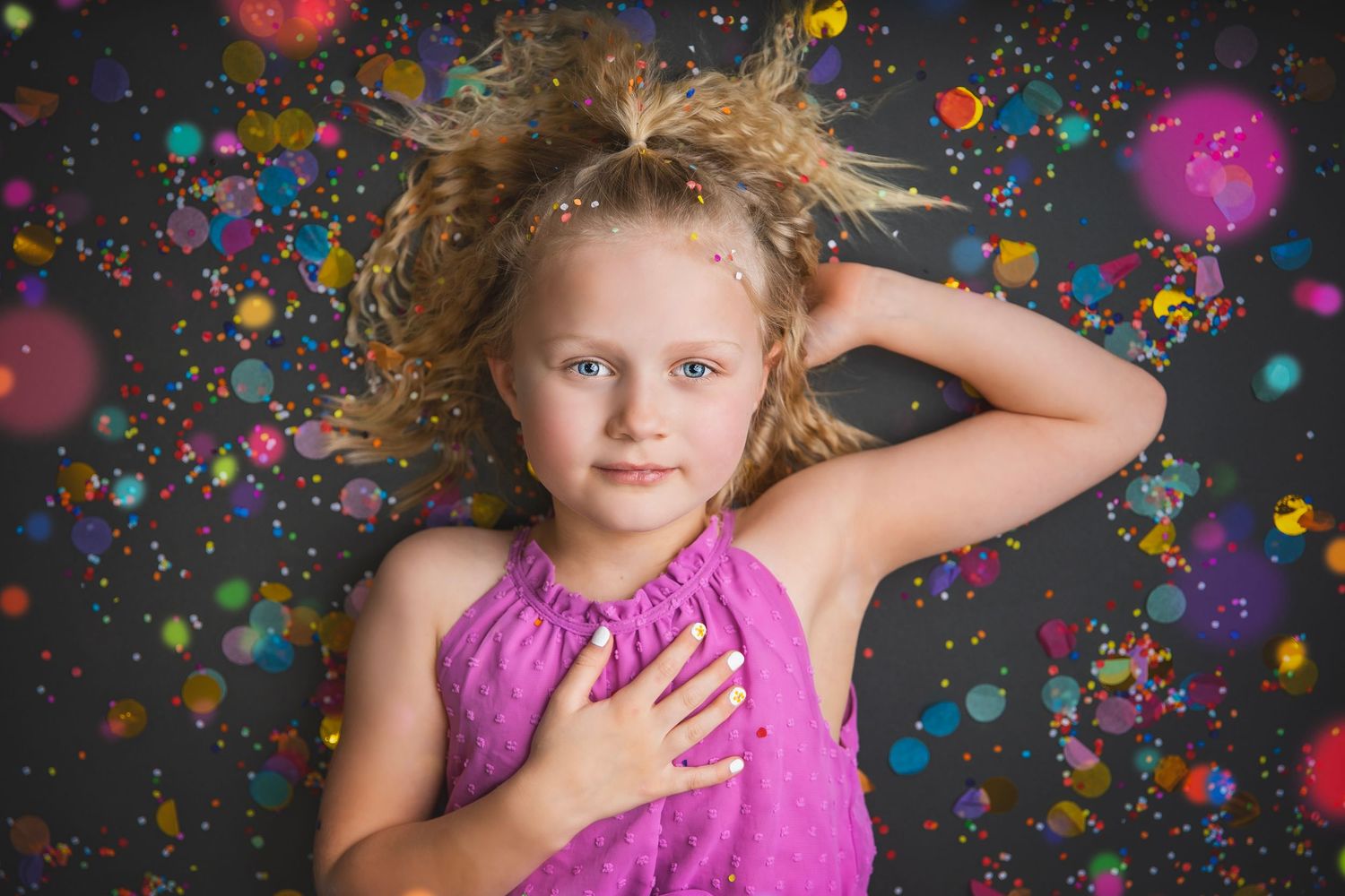 Blonde girl in pink dress lying down surrounded by colorful bokeh confetti on dark background.